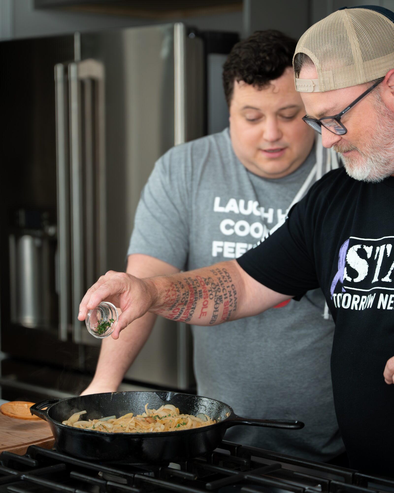 Two people cooking at a stovetop, with one pouring seasoning into a pan of food.