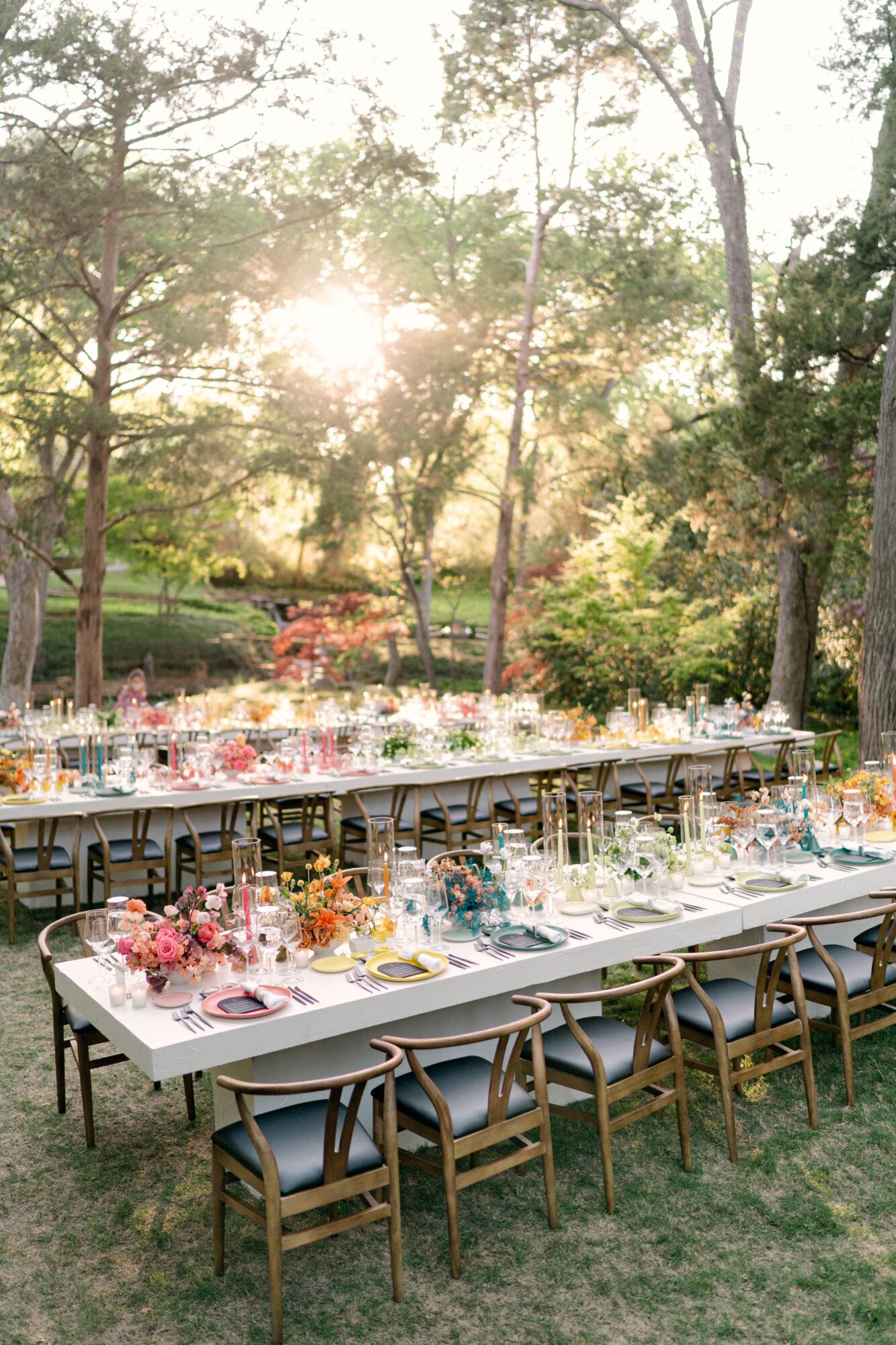 Long outdoor dining tables with place settings and flowers beneath trees in a sunlit garden.
