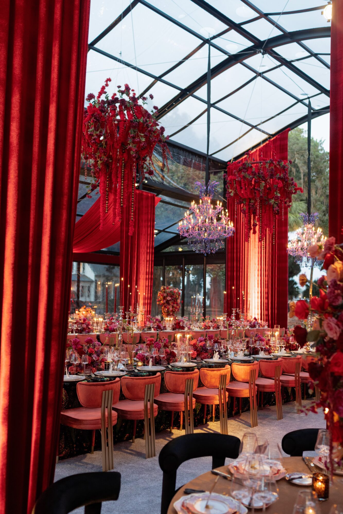 A dining room with red curtains, floral arrangements, chandeliers, and tables set with plates and glassware.