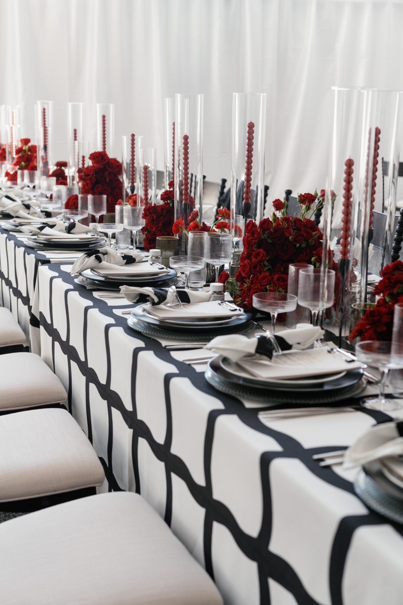 A long banquet table set with white plates, glassware, red floral arrangements, and white chairs.