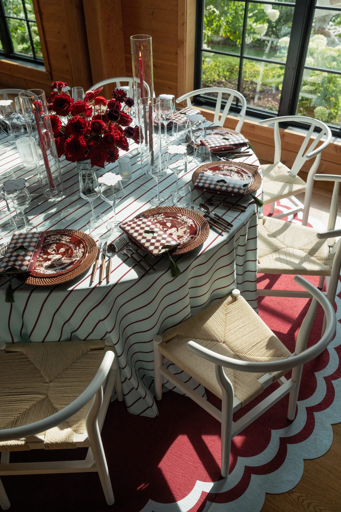 Dining table with patterned cloth, red flowers, place settings, and white chairs beside large windows.