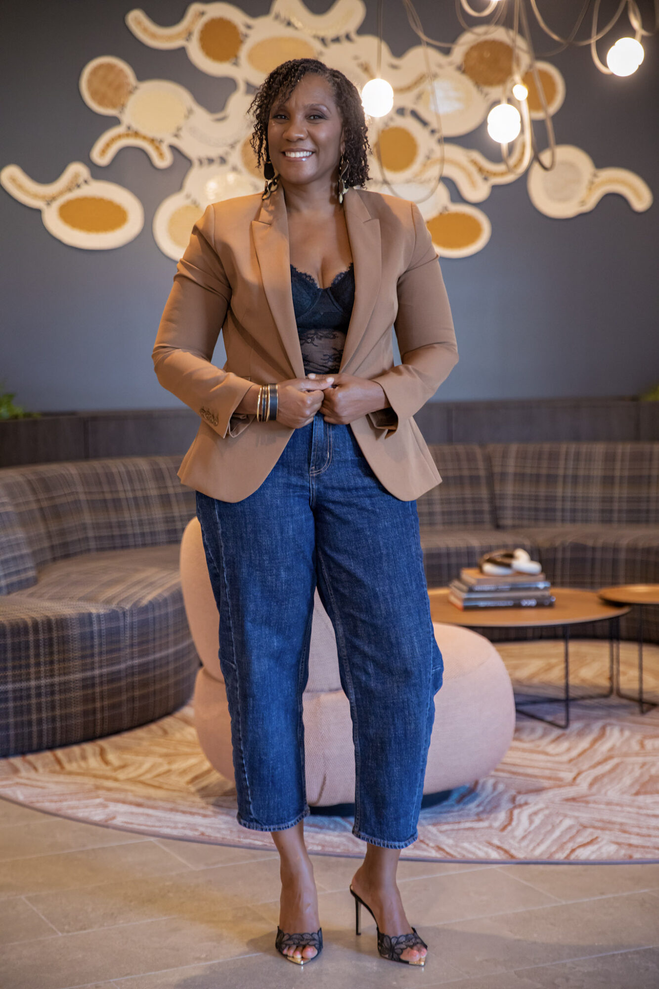 A person in a tan blazer and blue jeans stands indoors before a decorative wall sculpture and plaid seating.