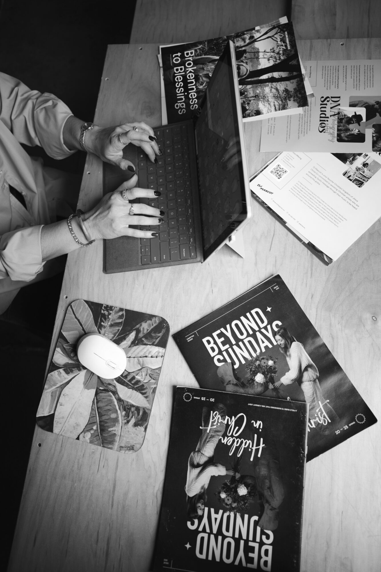 Person typing on a laptop at a table with books, papers, and a mouse.