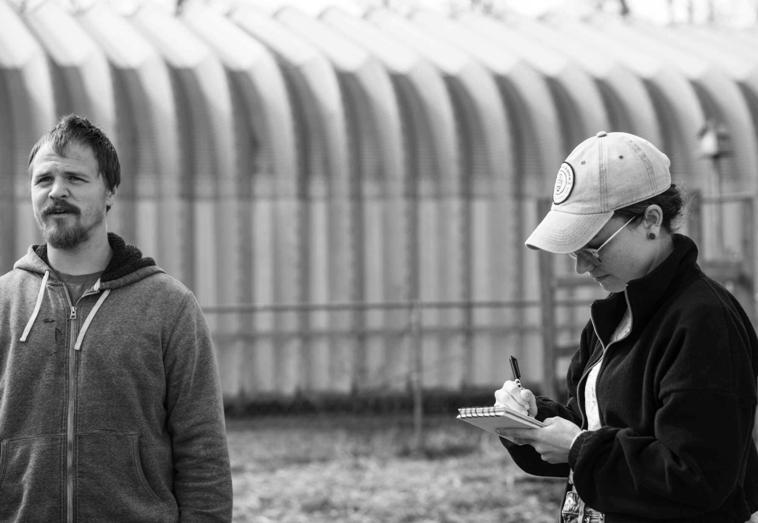 Two people stand beside a greenhouse; one looks toward the camera while the other writes in a notebook.