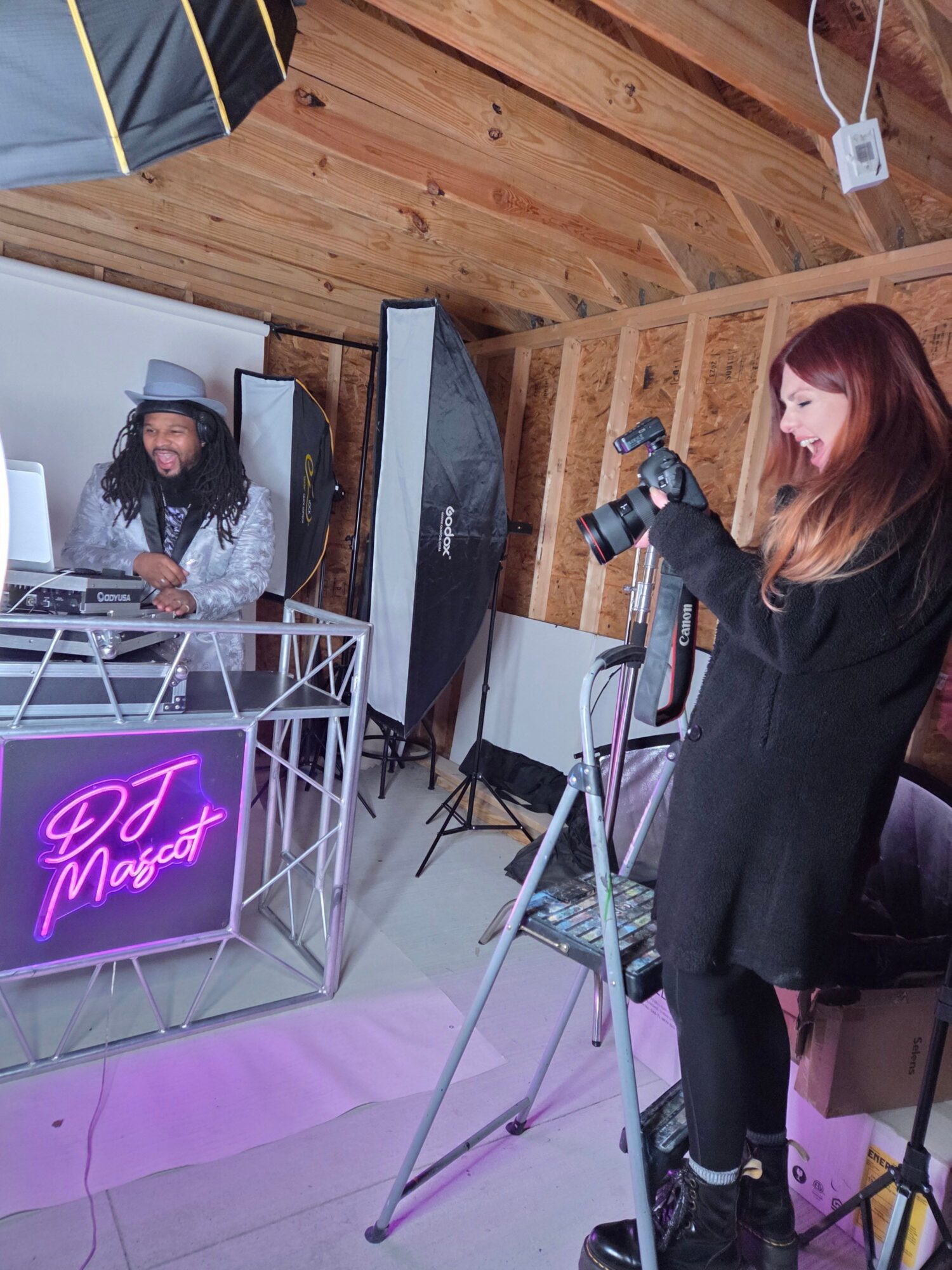 Two people stand near photography equipment and a glowing pink sign in a wood-paneled room.
