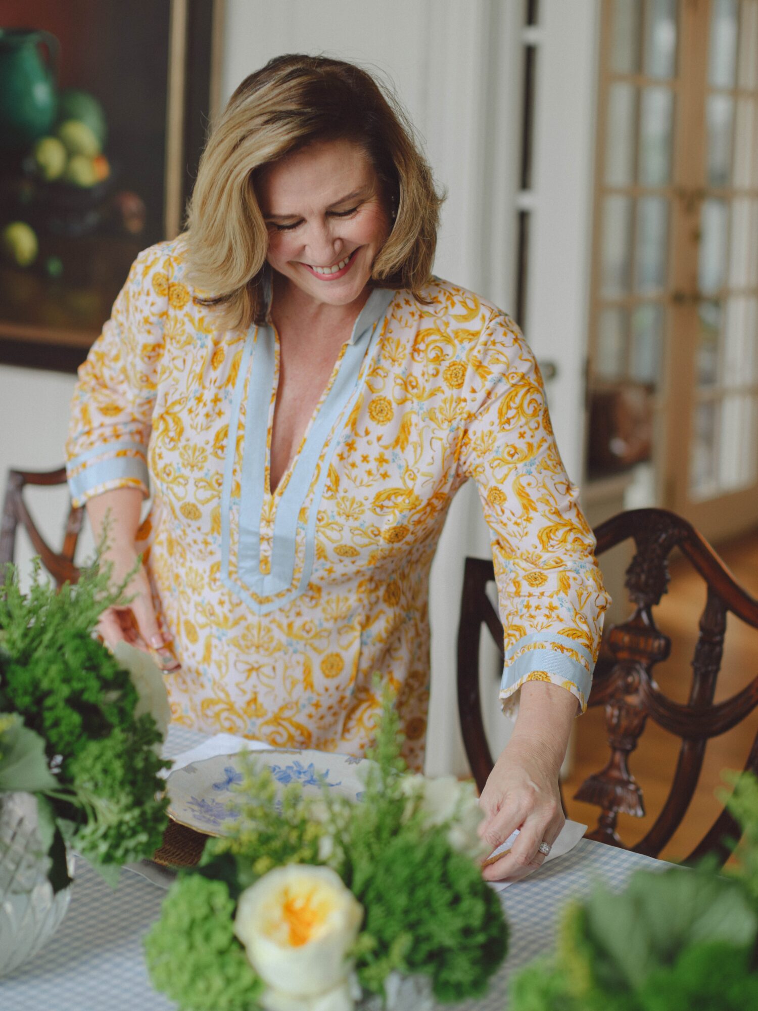 A woman in a yellow patterned blouse arranges greenery and flowers on a dining table.