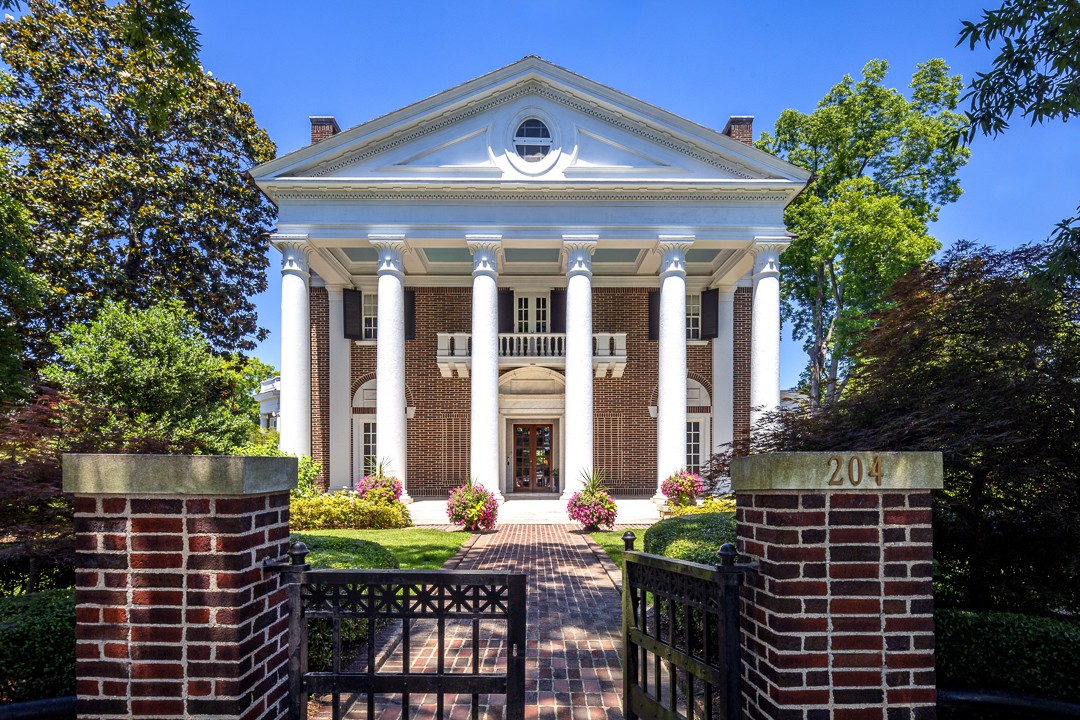 White-columned building behind an open black gate between brick pillars, with trees and shrubs under a blue sky.