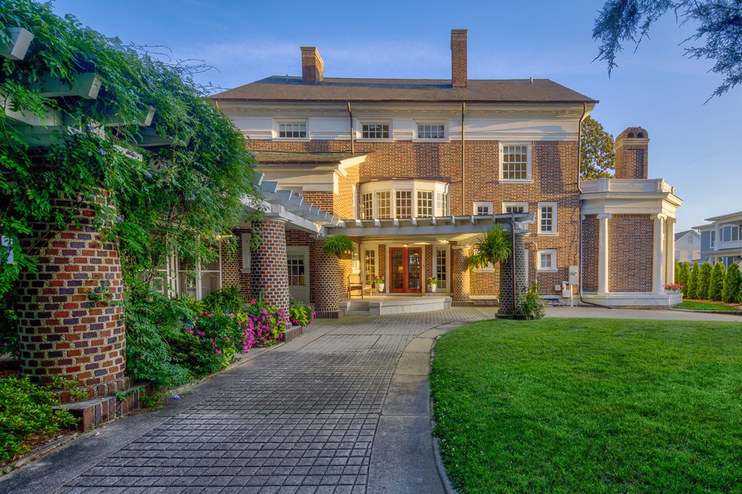 Brick house with a paved walkway, garden greenery, and a lawn under a blue sky.