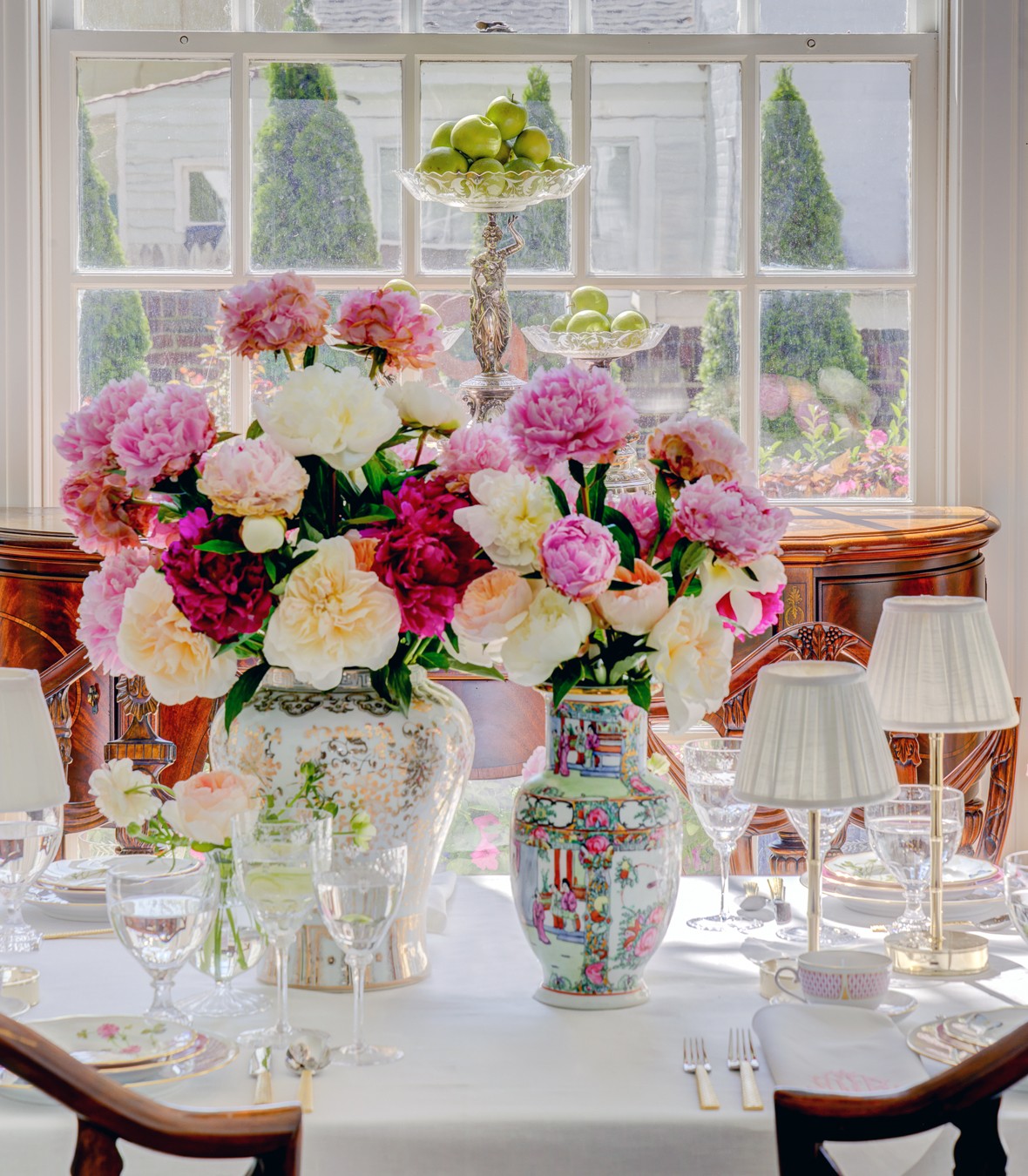 A dining table with colorful flower arrangements, glassware, and place settings in front of a large window.