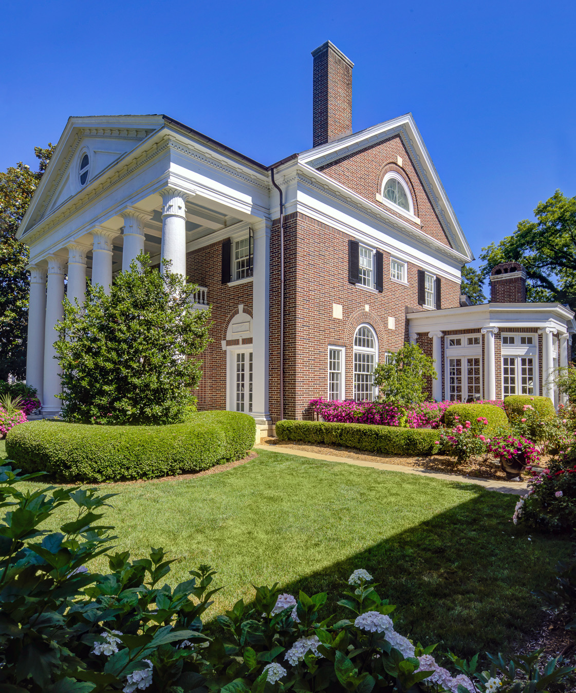 Red brick house with white columns, a tall chimney, green lawn, and landscaped flower beds.