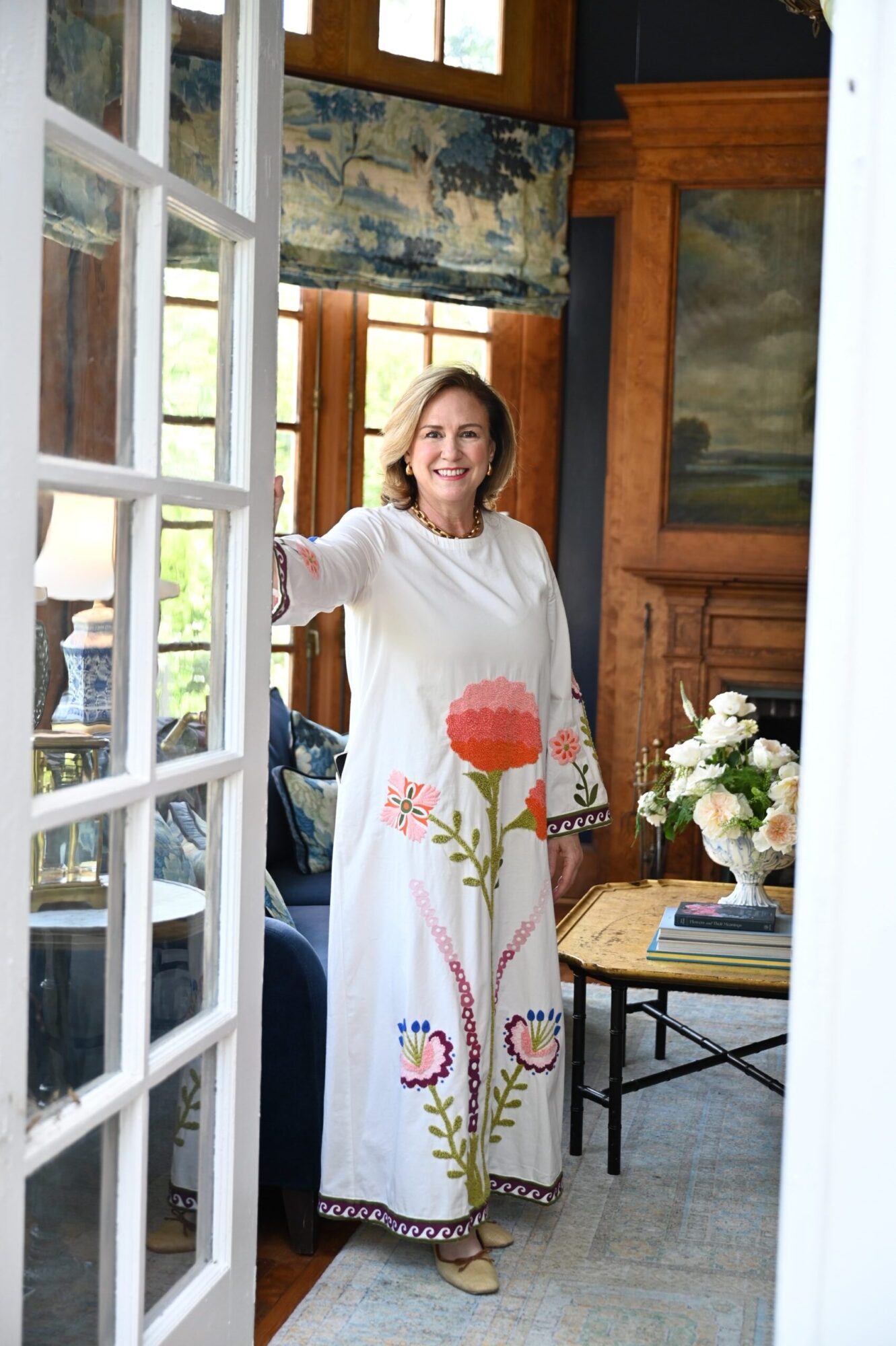 A person in a white floral shirt stands beside an open window in a room with wooden doors and flowers.