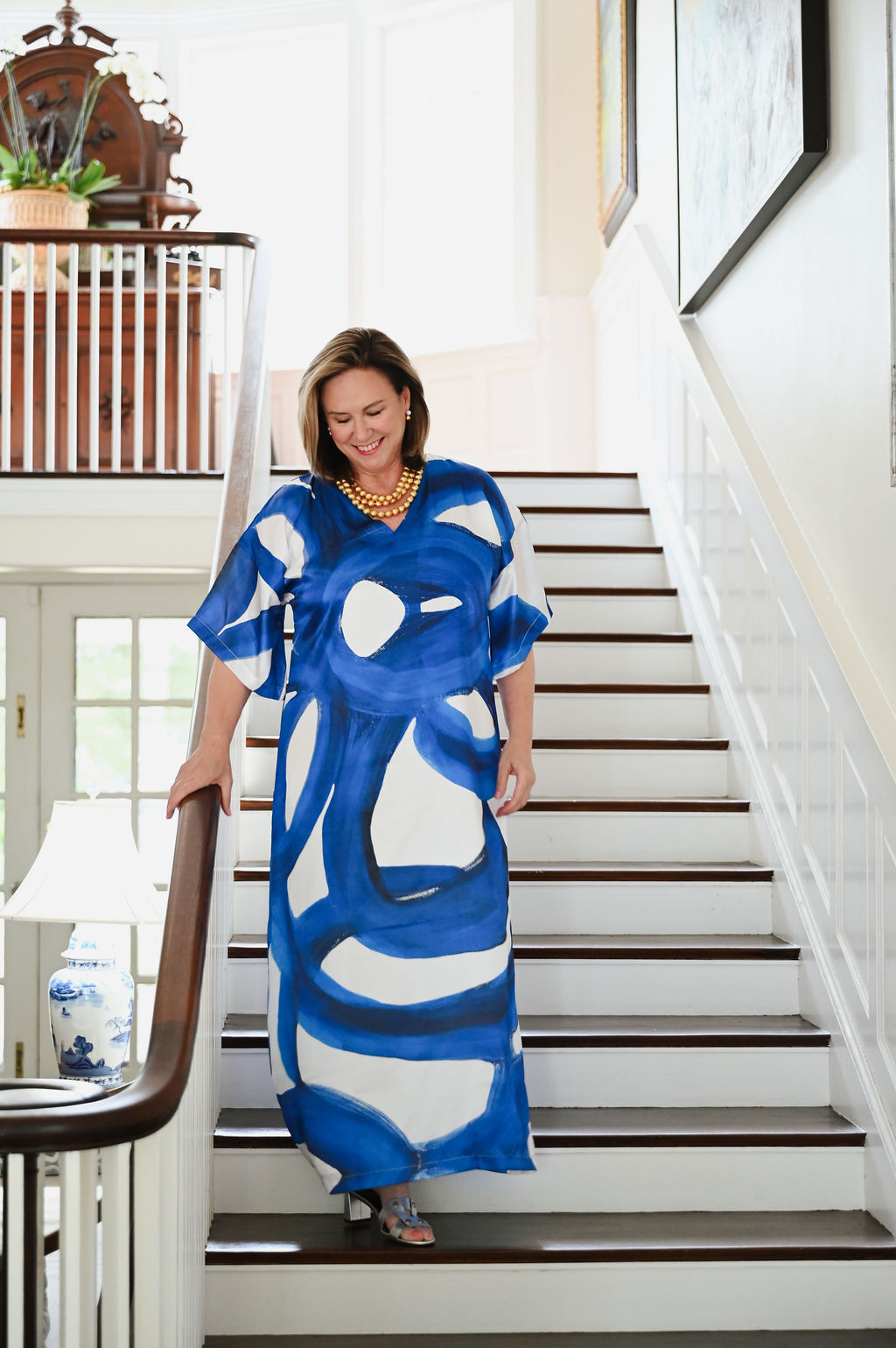 A person in a blue patterned dress descends a staircase with a railing and framed pictures.