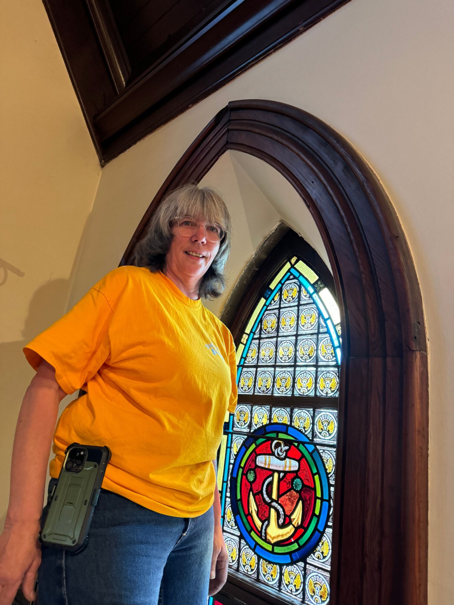A person in an orange shirt stands beside a stained-glass window inside a building.