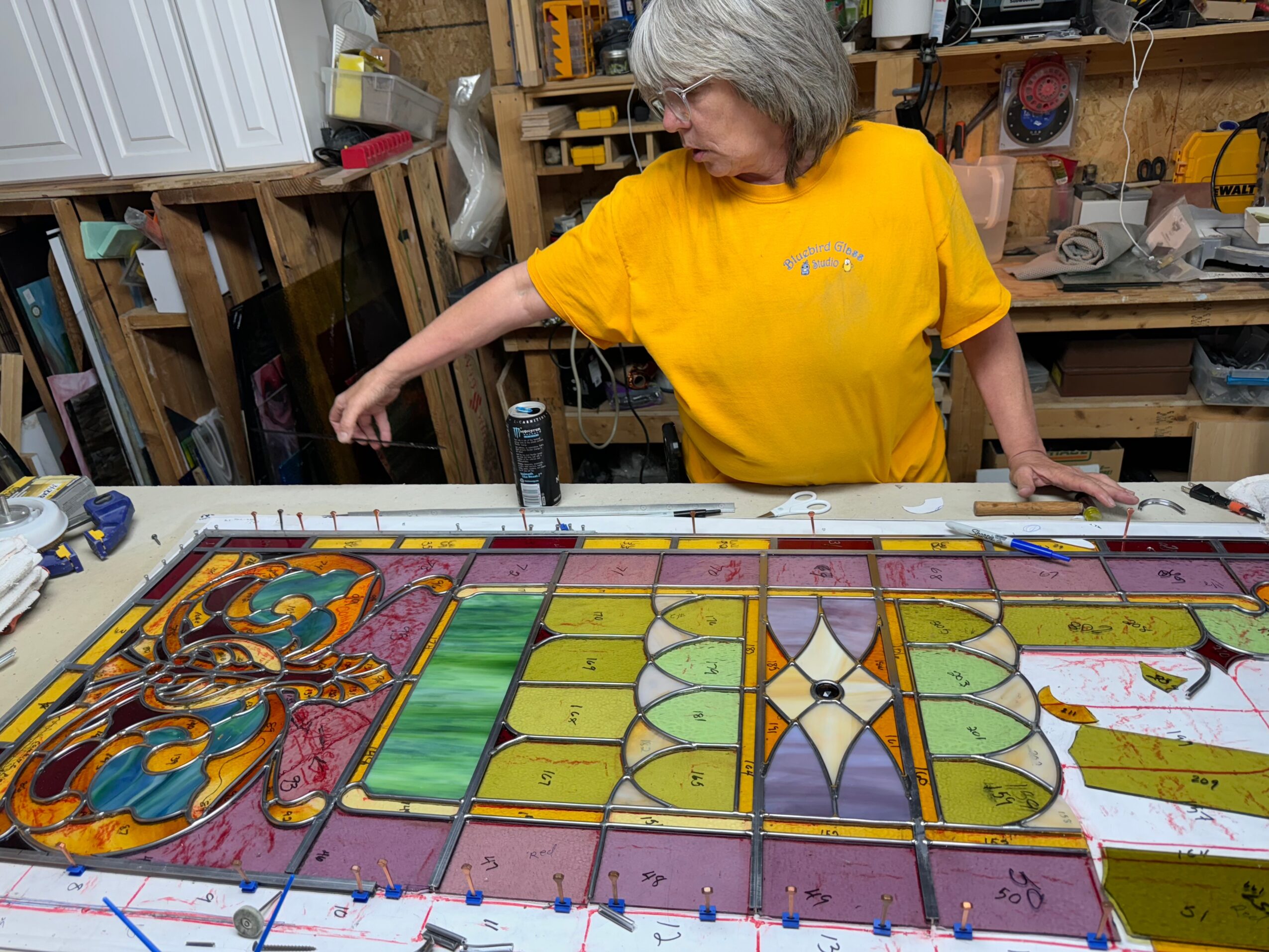 Person in a yellow shirt working over a colorful stained-glass panel in a workshop.