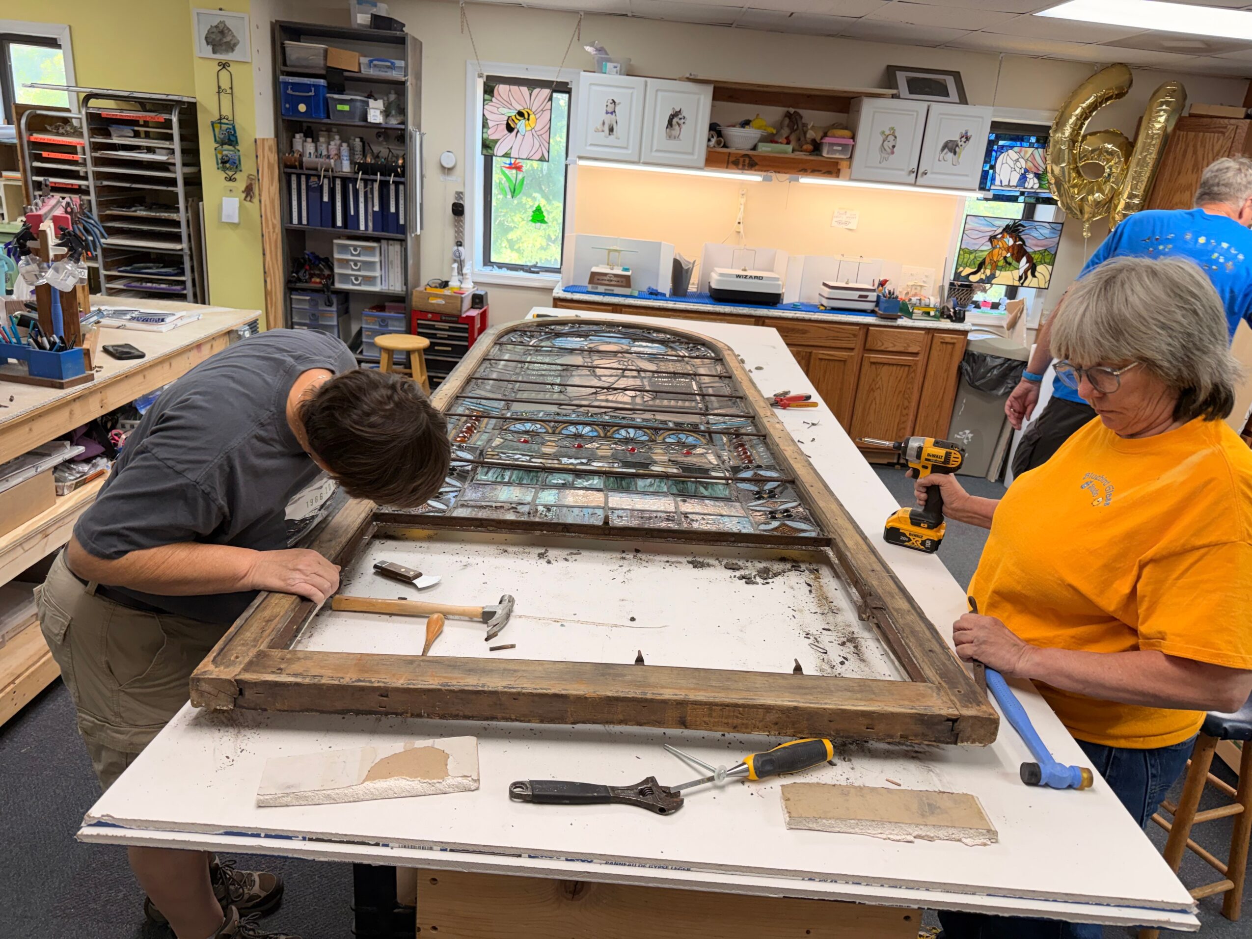 Two people work on a large wooden frame at a workshop table, surrounded by tools and shelves.