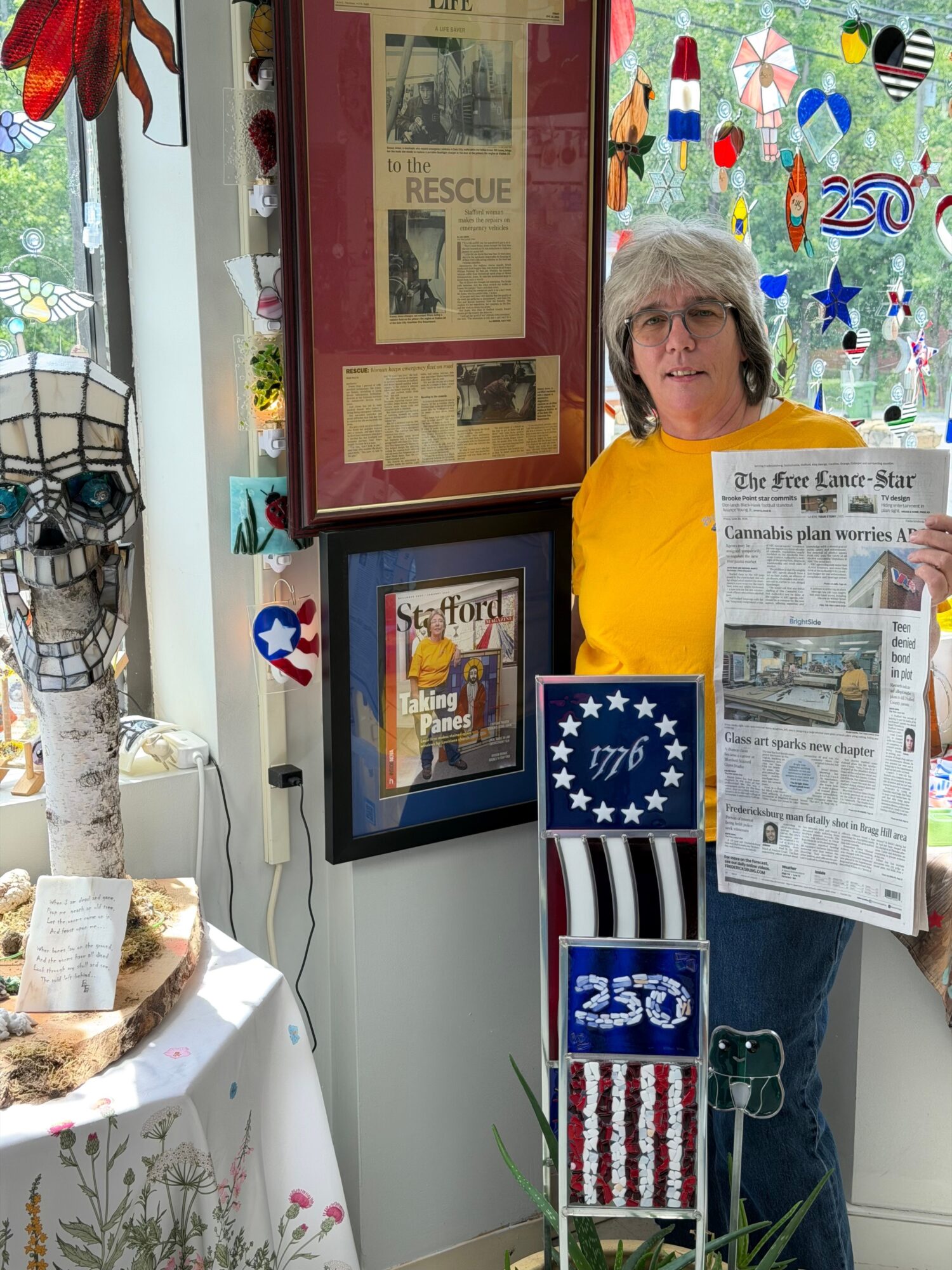 Person holding a newspaper beside framed newspaper clippings, a U.S. flag display, and colorful artwork.