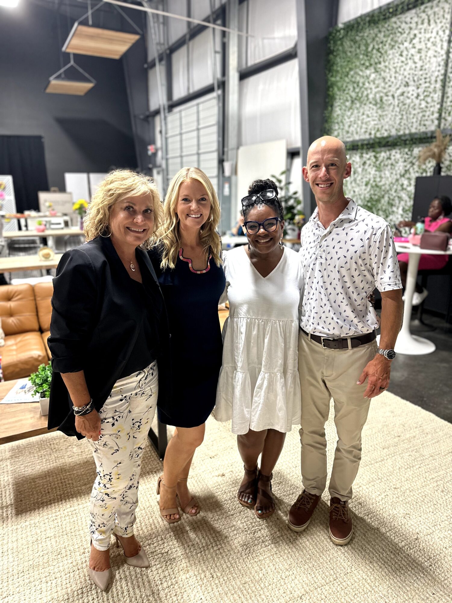 Four people stand together in a bright indoor workspace with tables, chairs, and a green wall.