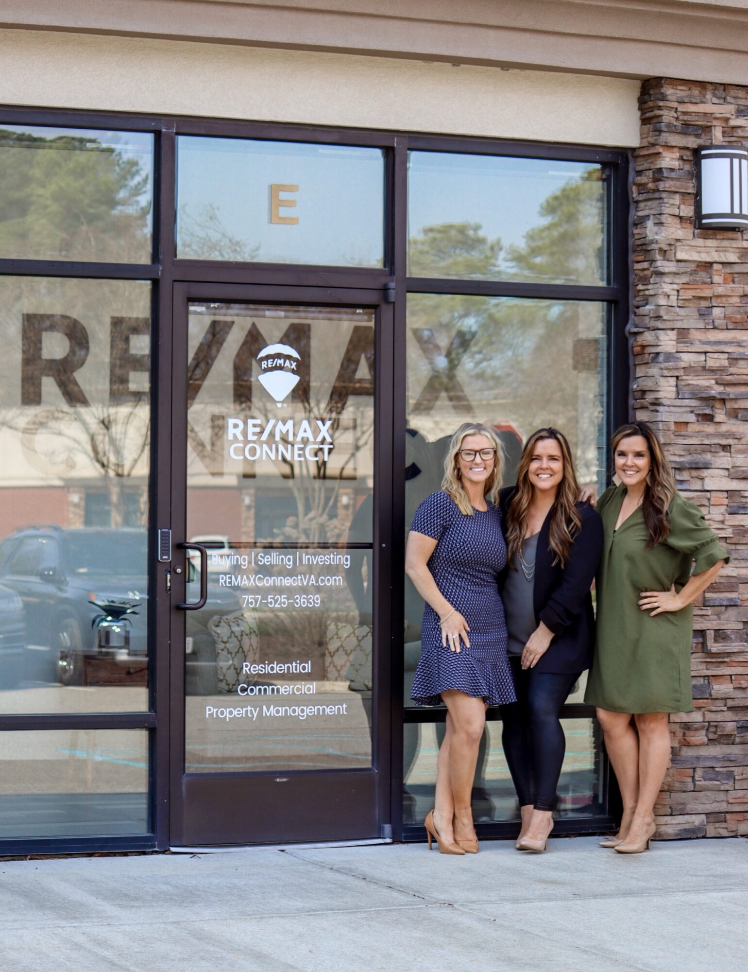 Three people stand outside a RE/MAX real estate office with glass doors and stone siding.
