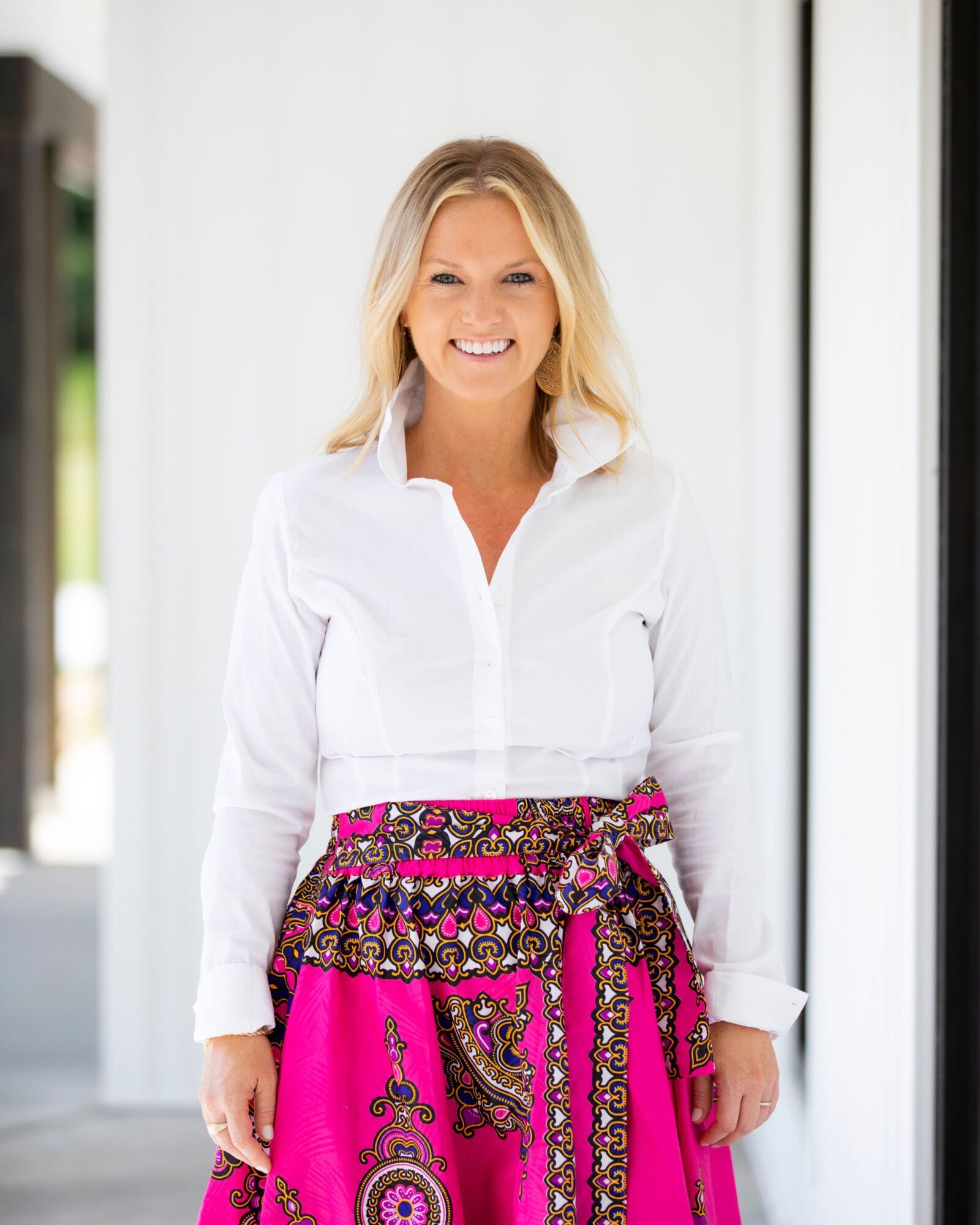 A smiling person in a white blouse and bright pink patterned skirt stands in a bright interior.
