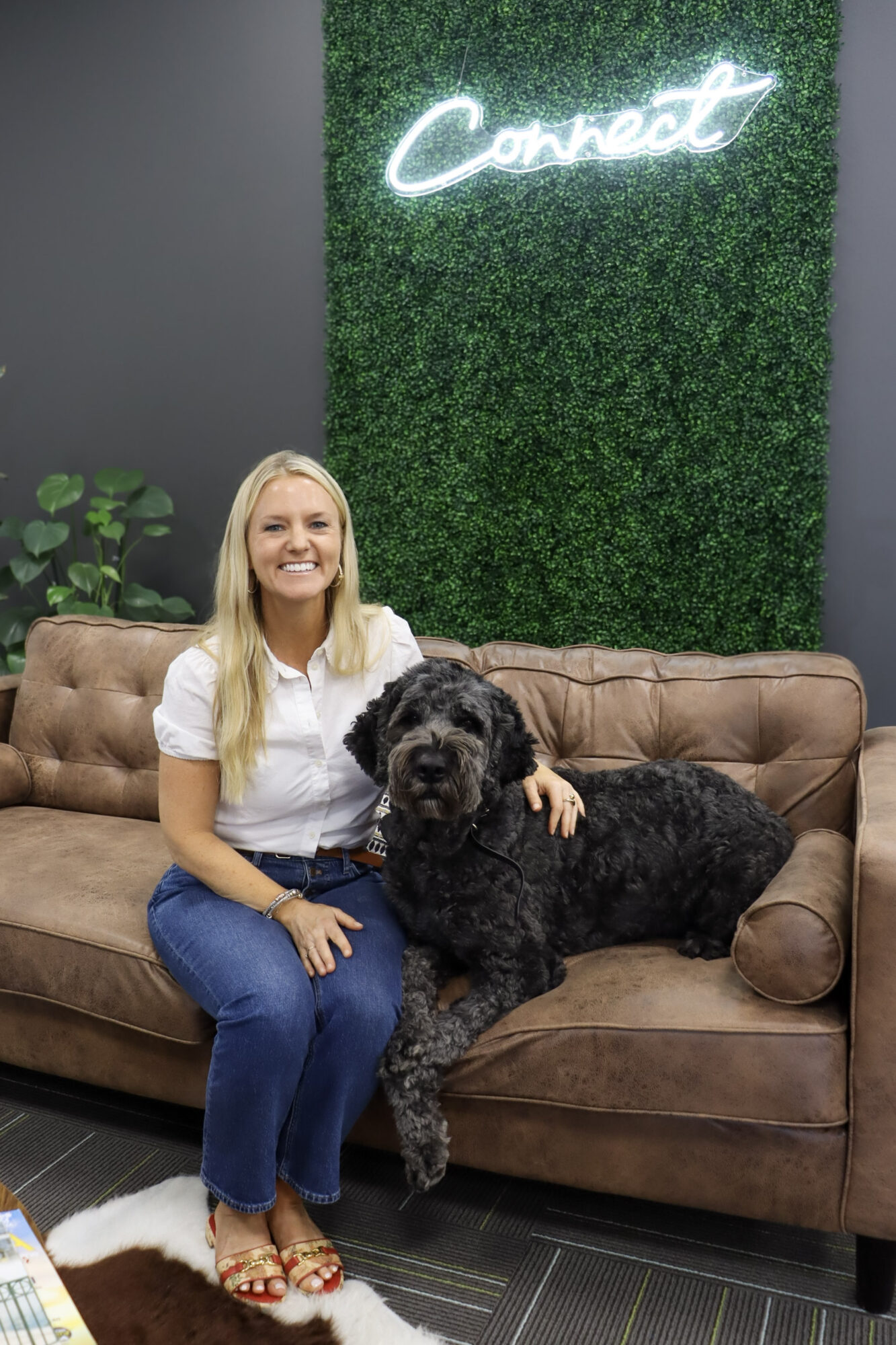 A woman sits on a brown couch beside a large black dog, with a green wall and illuminated sign behind them.