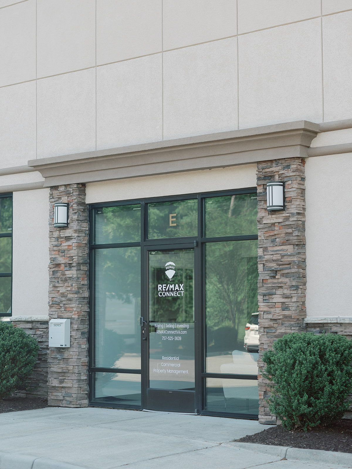 Glass entrance framed by stone columns, with a RE/MAX sign on the door and shrubs on either side.