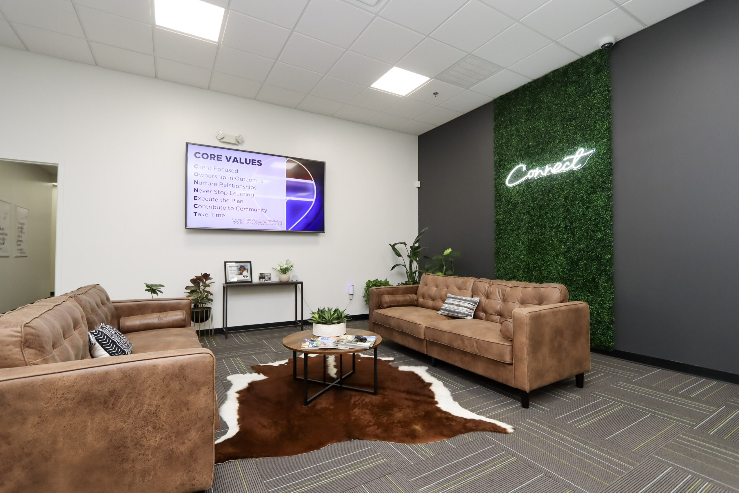Office lounge with brown sofas, a coffee table, rug, wall-mounted presentation screen, and green feature wall.