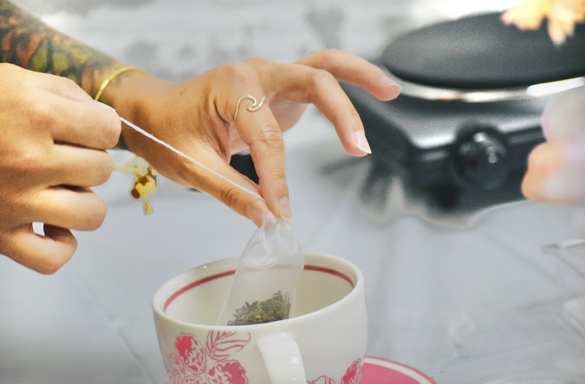 Hands place a tea bag into a floral cup beside a dark kettle.
