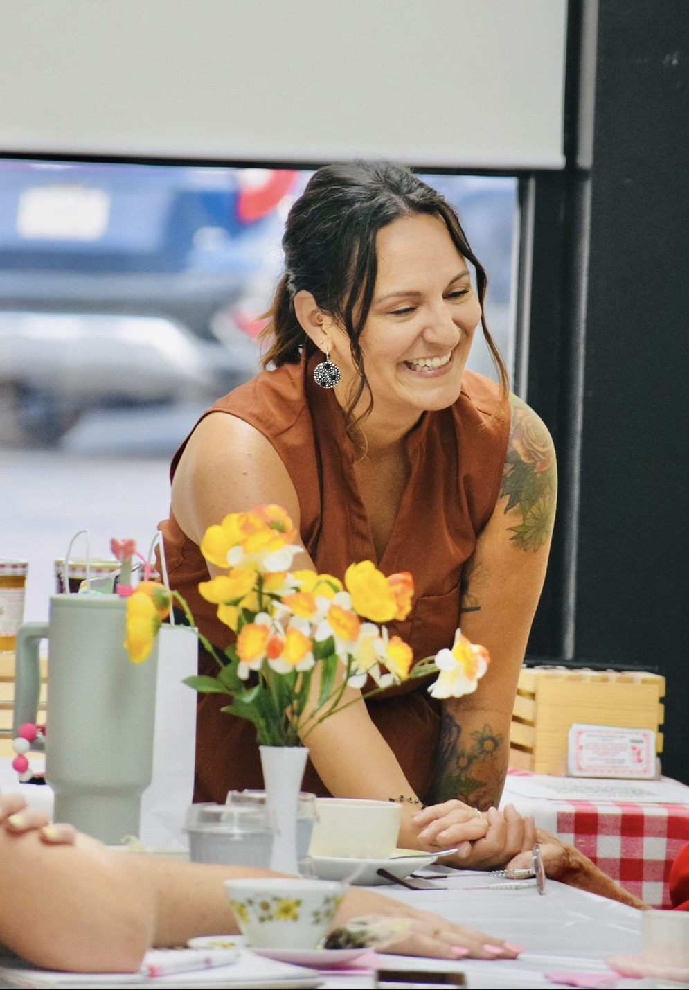 A woman leans over a table with yellow flowers, cups, and a red-checkered cloth.
