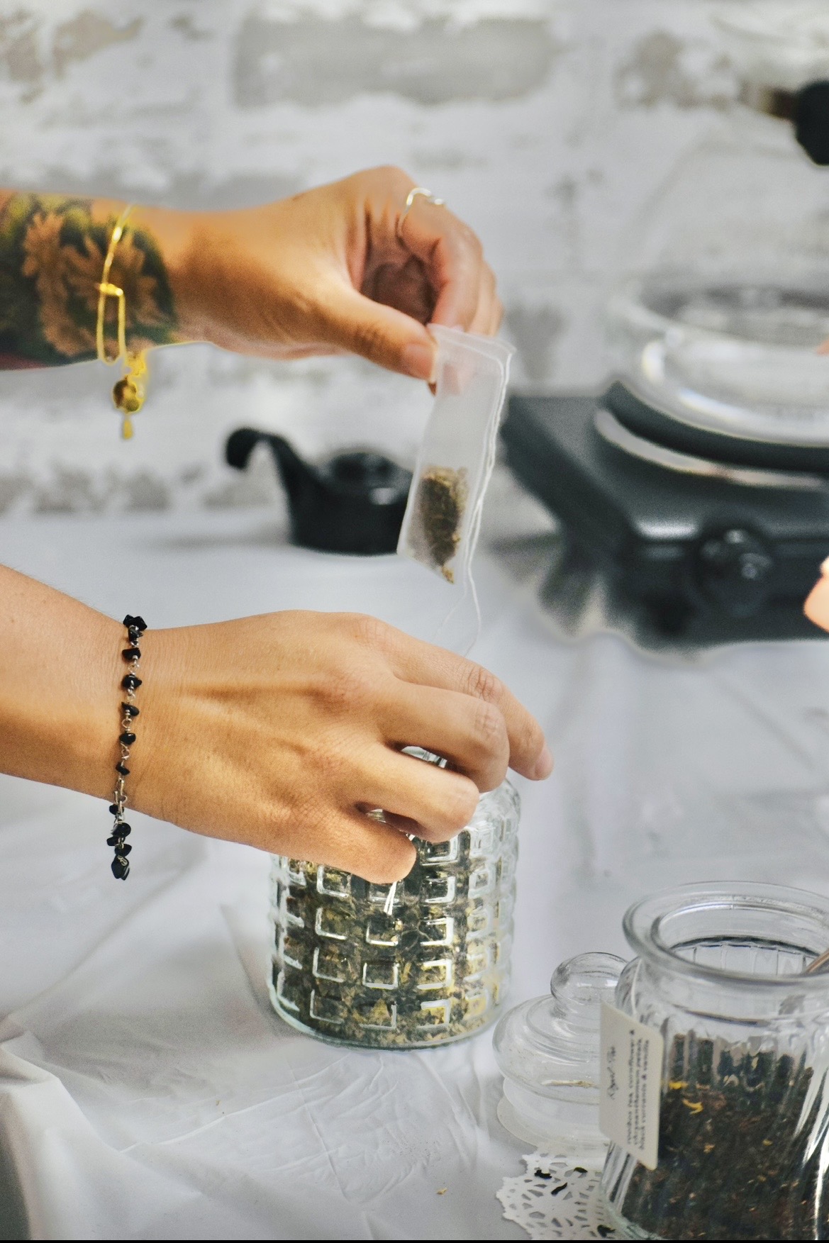 Hands pour dried herbs from a small packet into a glass jar beside a bowl of herbs.