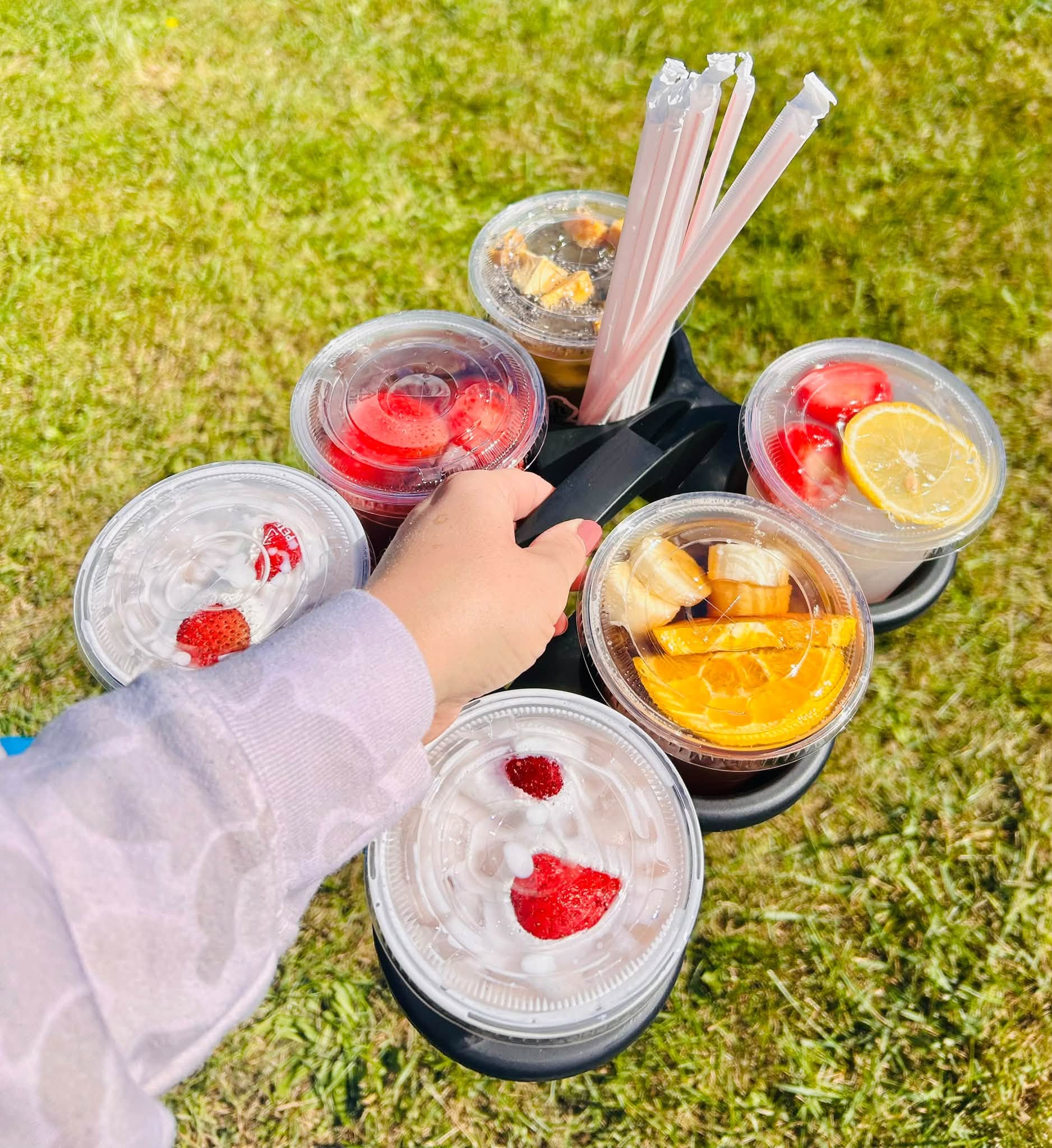 A hand holds several plastic cups of colorful fruit and dessert toppings with wrapped utensils on grass.