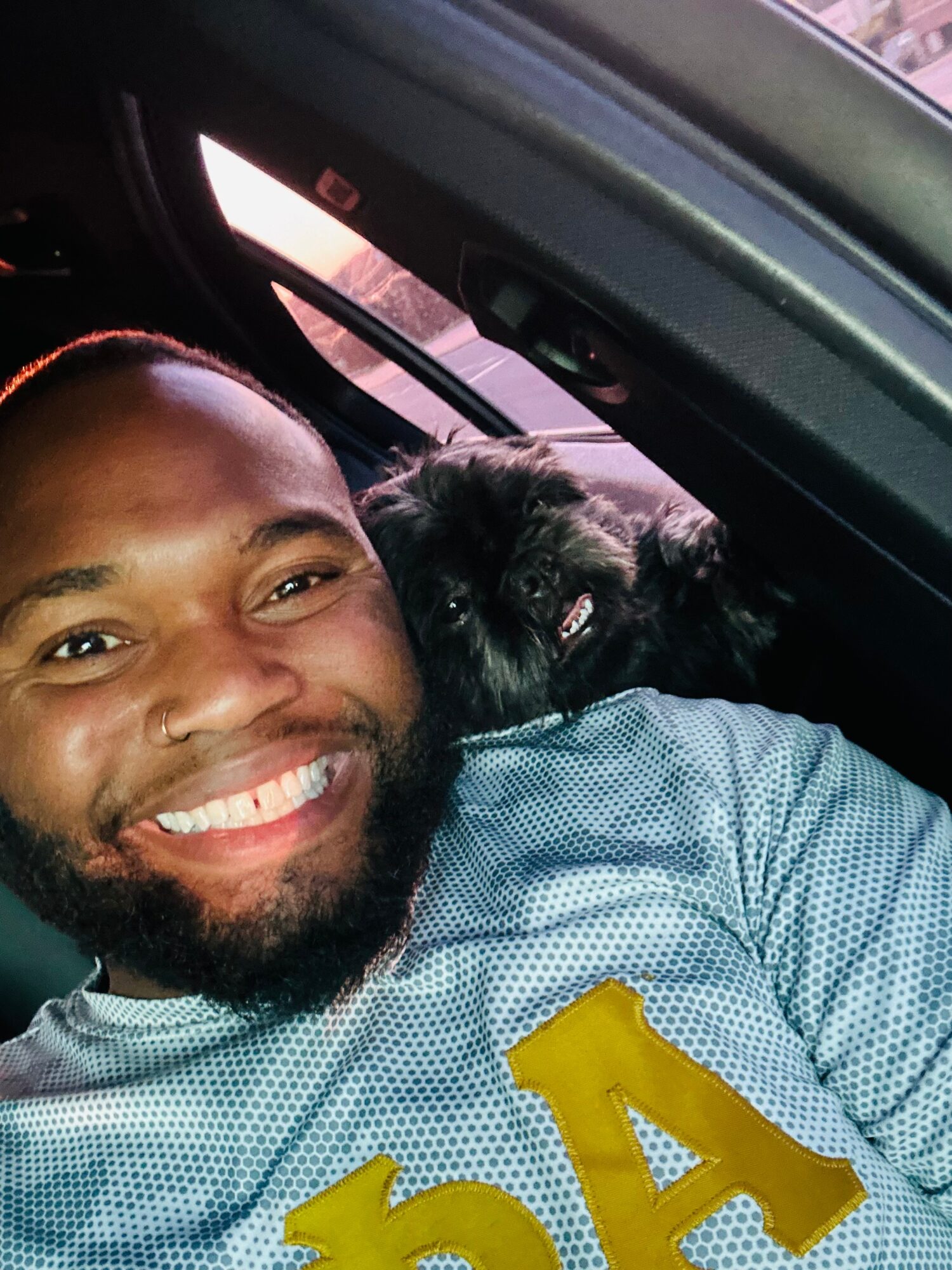 Smiling person in a blue patterned jersey beside a black dog inside a car.