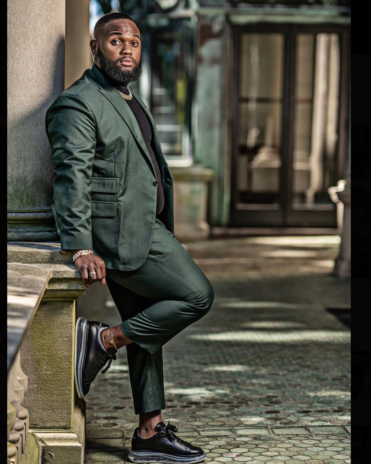 A person in a dark green suit leans against a stone railing outside a glass doorway.