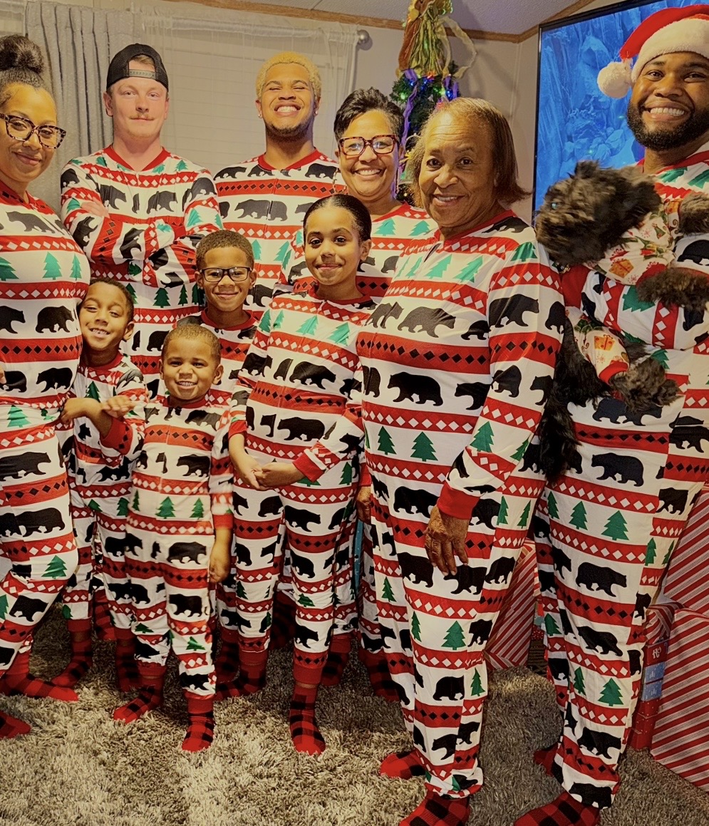 A group of adults and children in matching red, green, and white holiday pajamas pose beside a Christmas tree.