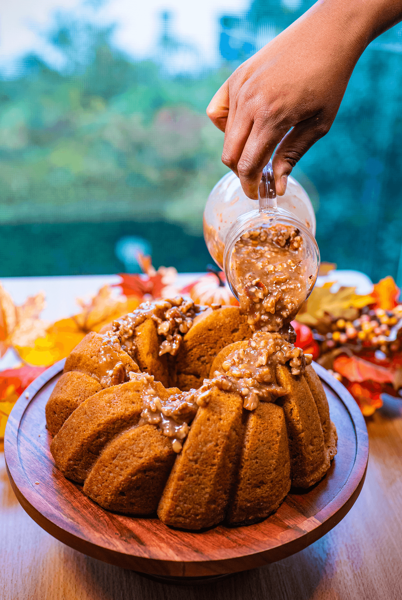 A hand pours sauce from a small glass pitcher over a ring-shaped cake on a wooden platter.
