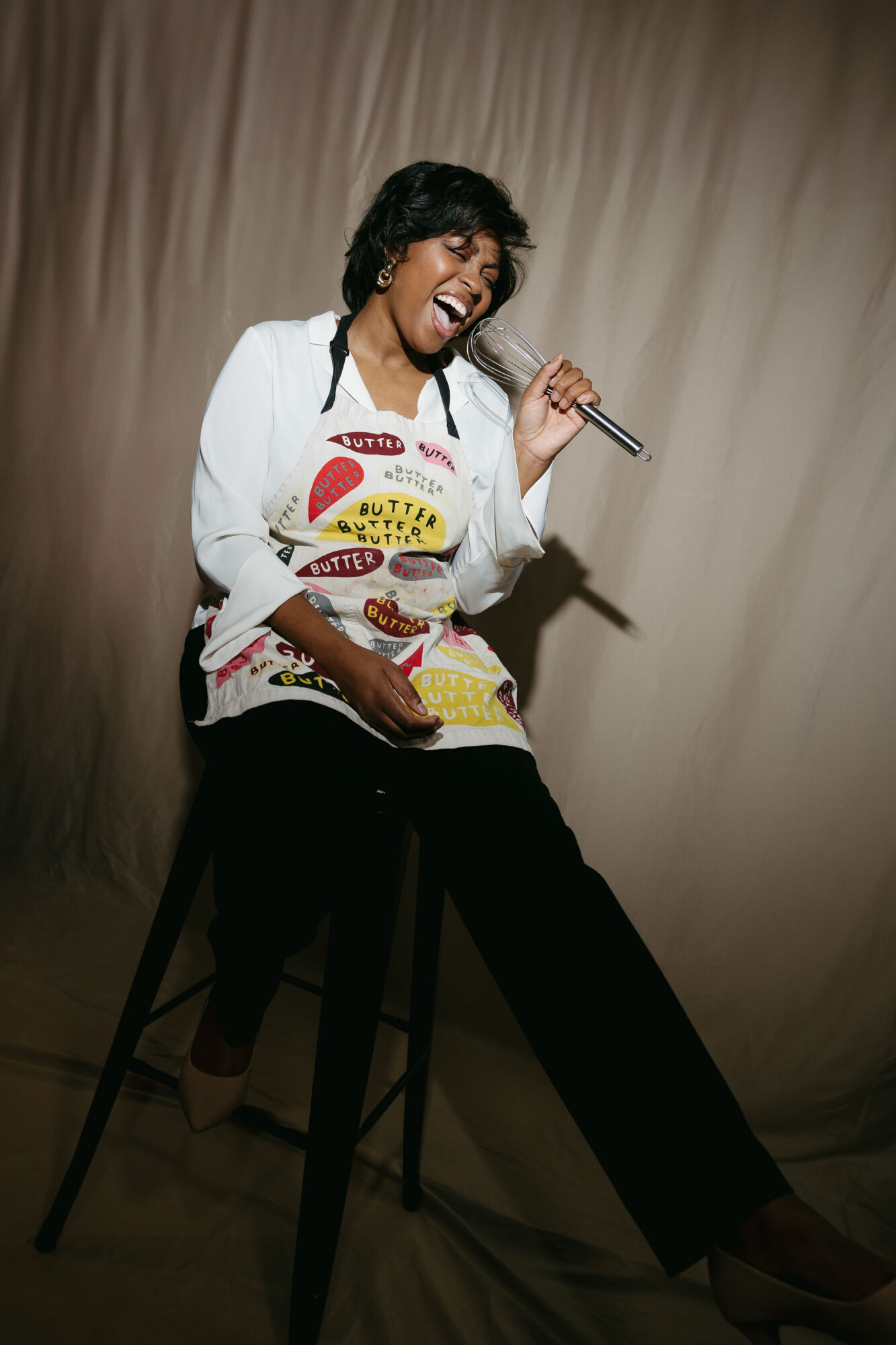 A person in a white jacket and colorful apron sits on a stool, holding a microphone and singing.