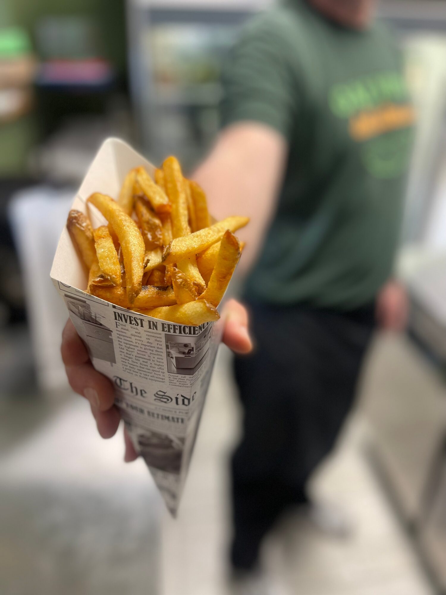 A person holds a paper cone filled with fries toward the camera.