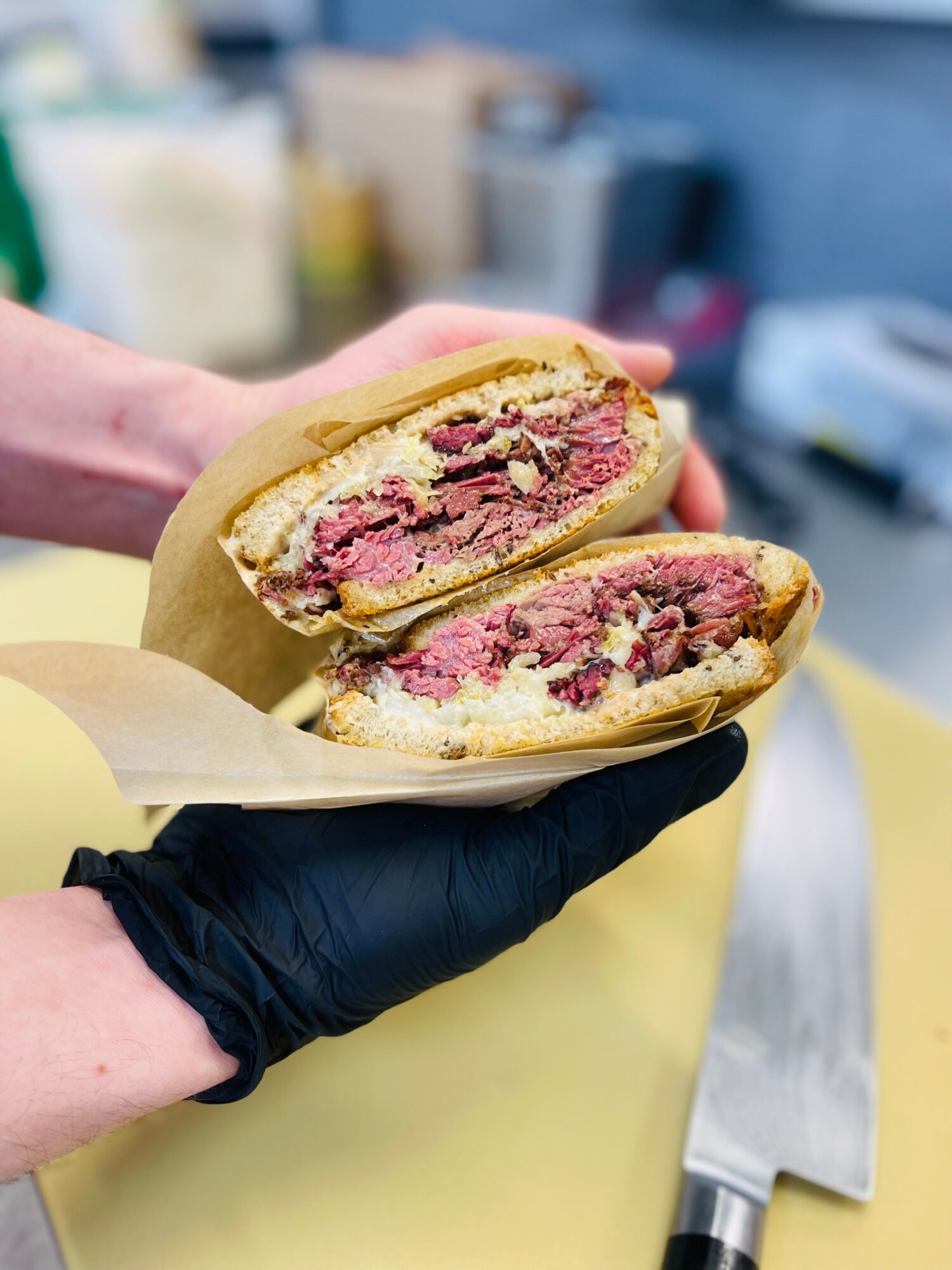 A gloved hand holds a split sandwich filled with sliced meat and other ingredients over a work surface.