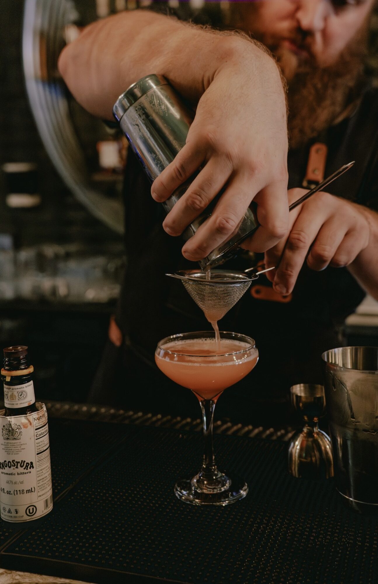 Bartender pours a drink through a strainer into a stemmed glass behind a bar.