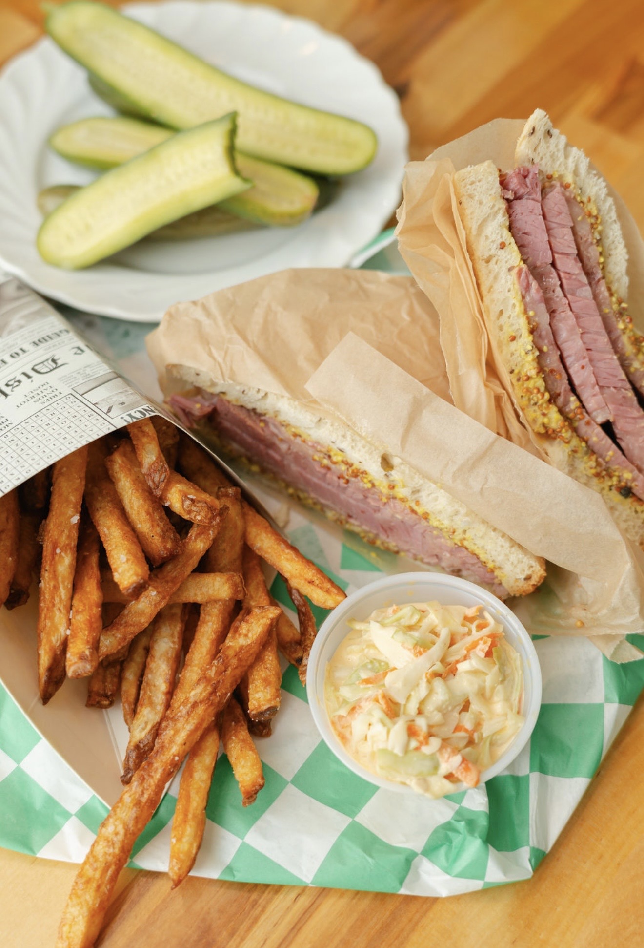 A plate of cucumber slices, two sandwich portions, seasoned fries, and a small cup of coleslaw on a checkered liner.