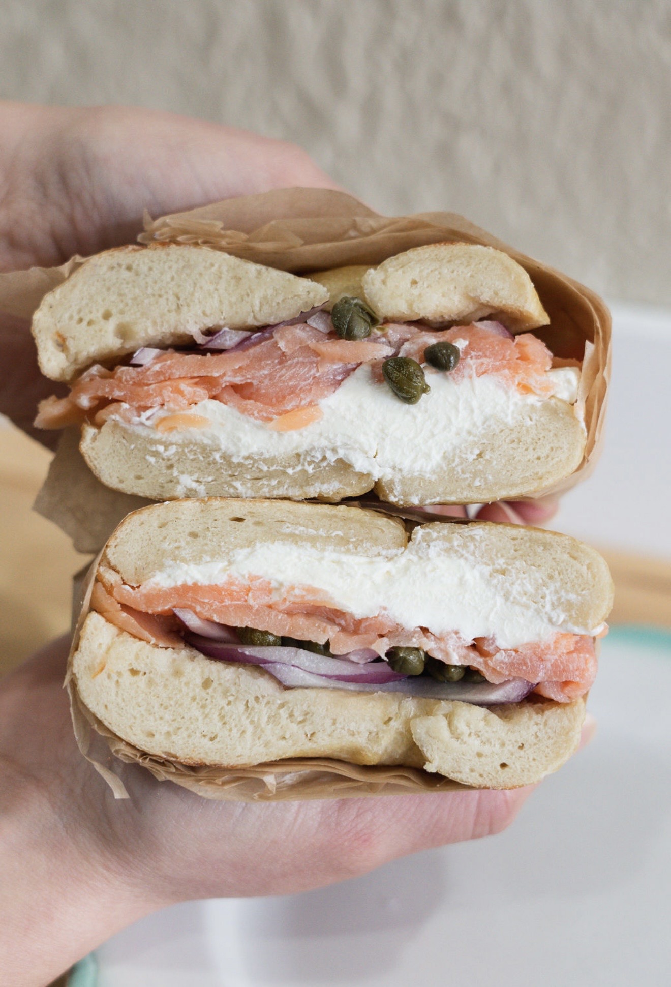 A hand holds a cut bagel sandwich layered with cream cheese, salmon, onion, and capers.