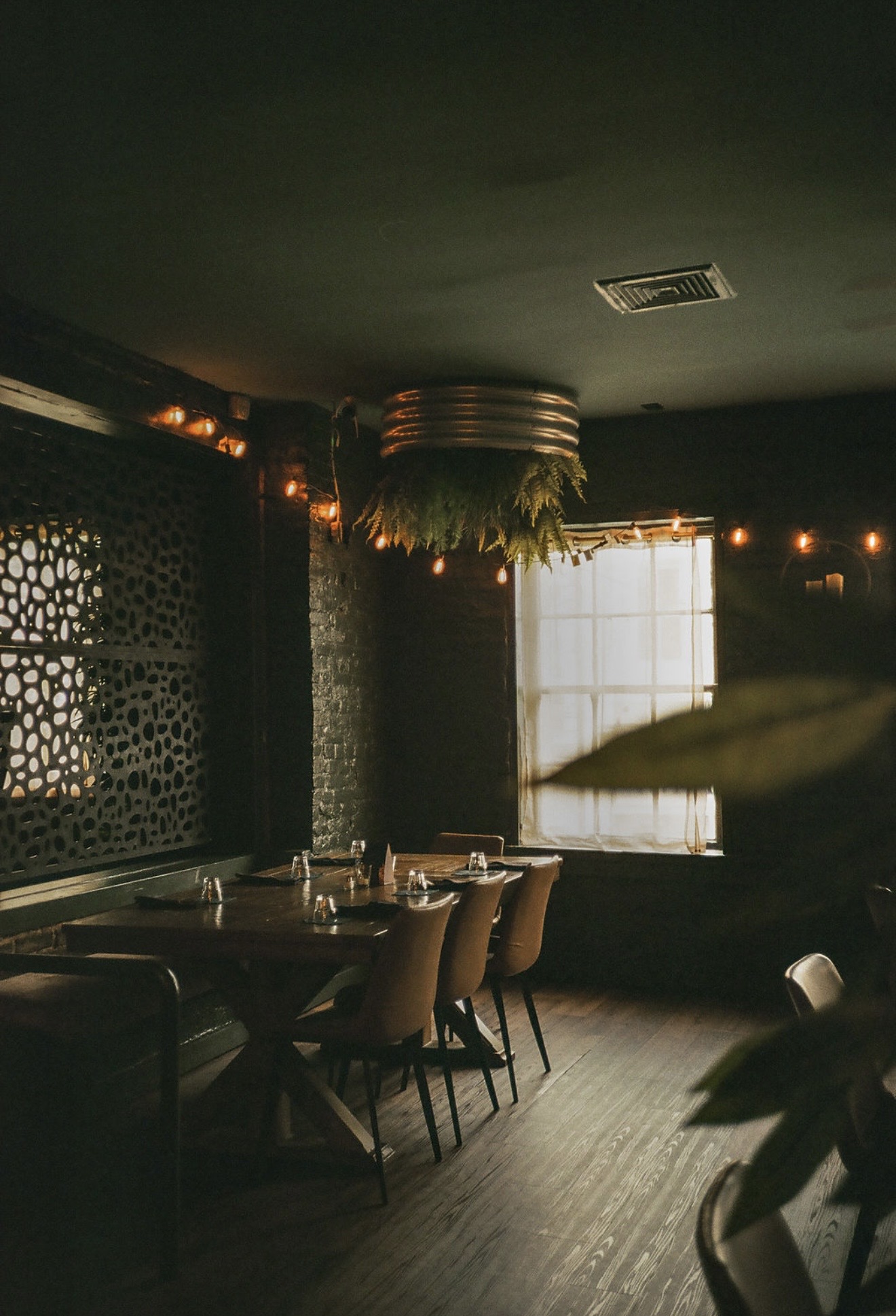 Dimly lit restaurant with tables, chairs, patterned wall panels, and a bright window.