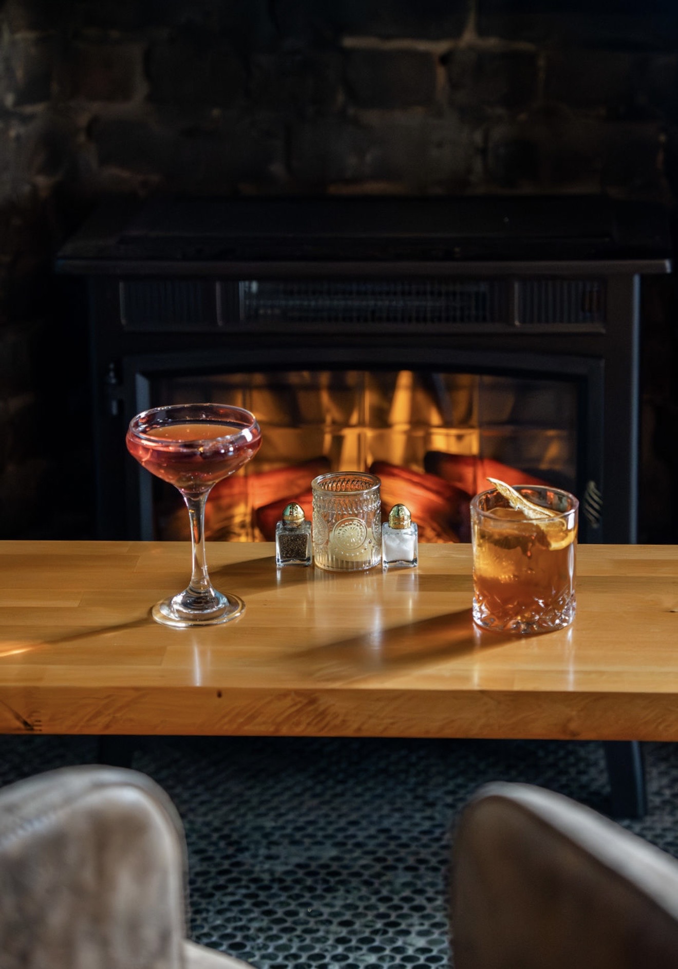 Two cocktails sit on a wooden table in front of a lit fireplace, with upholstered chairs in the foreground.