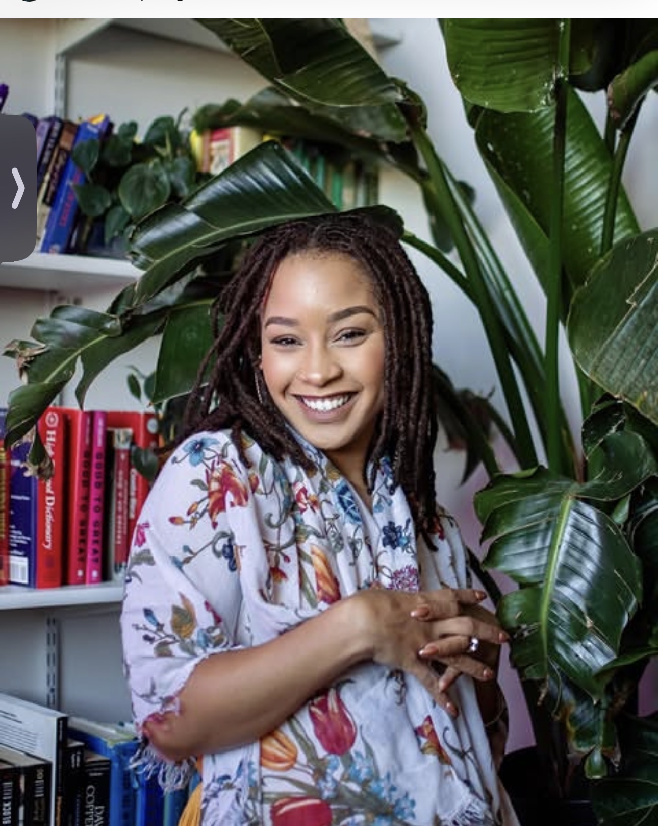 Smiling person with long locs stands among large leafy plants and bookshelves, wearing a floral robe.