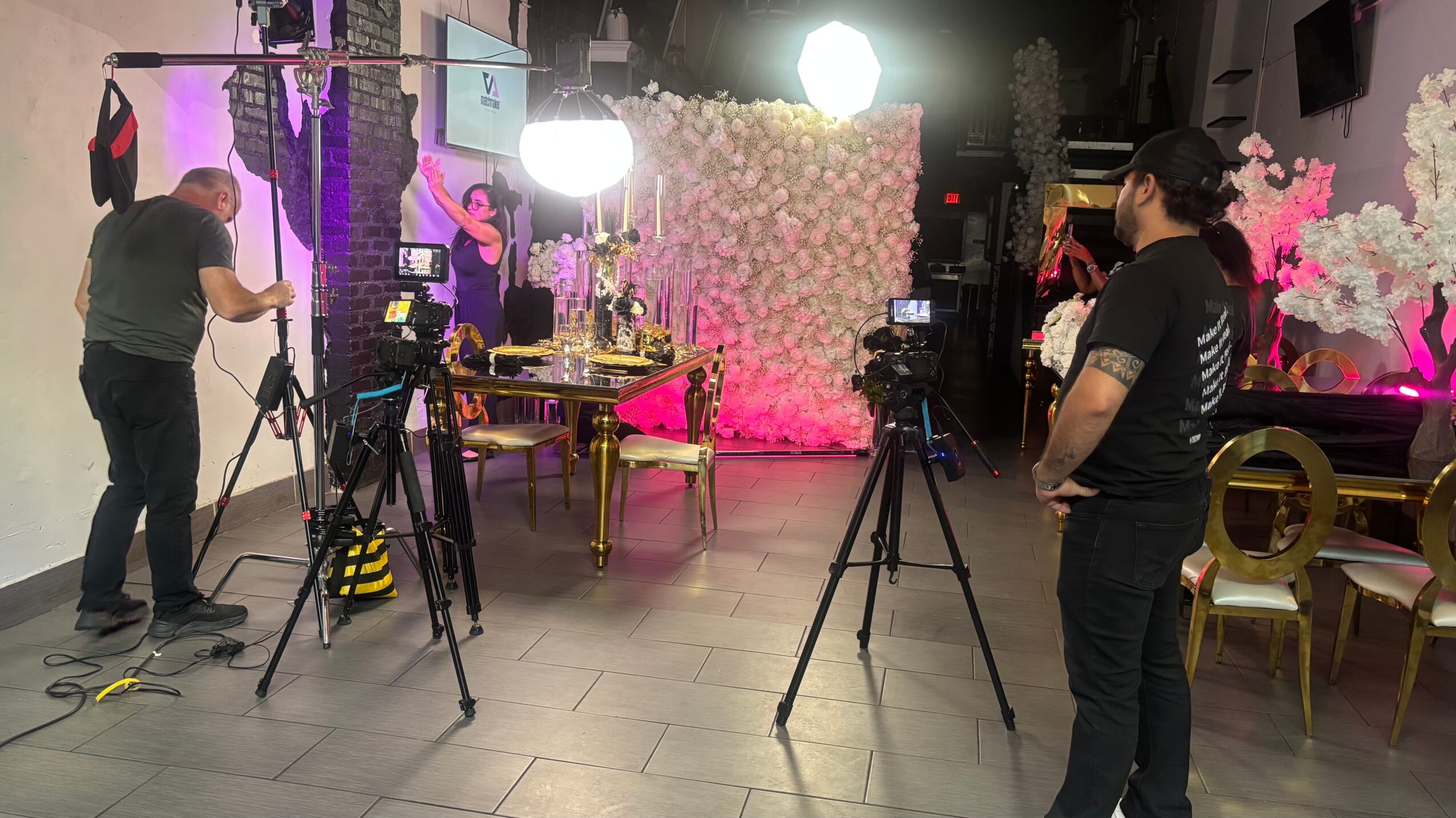 Photography studio with people, cameras, lighting equipment, a floral backdrop, and chairs.