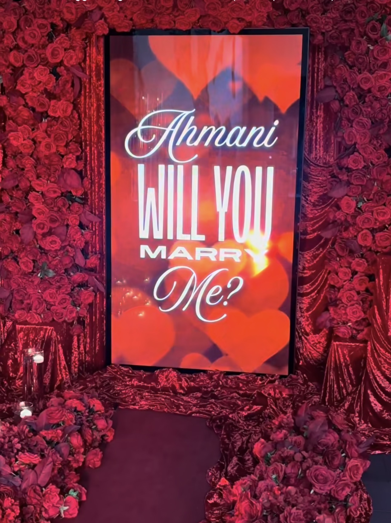 A framed proposal sign reading “Ahnani, will you marry me?” surrounded by red flowers and candles.