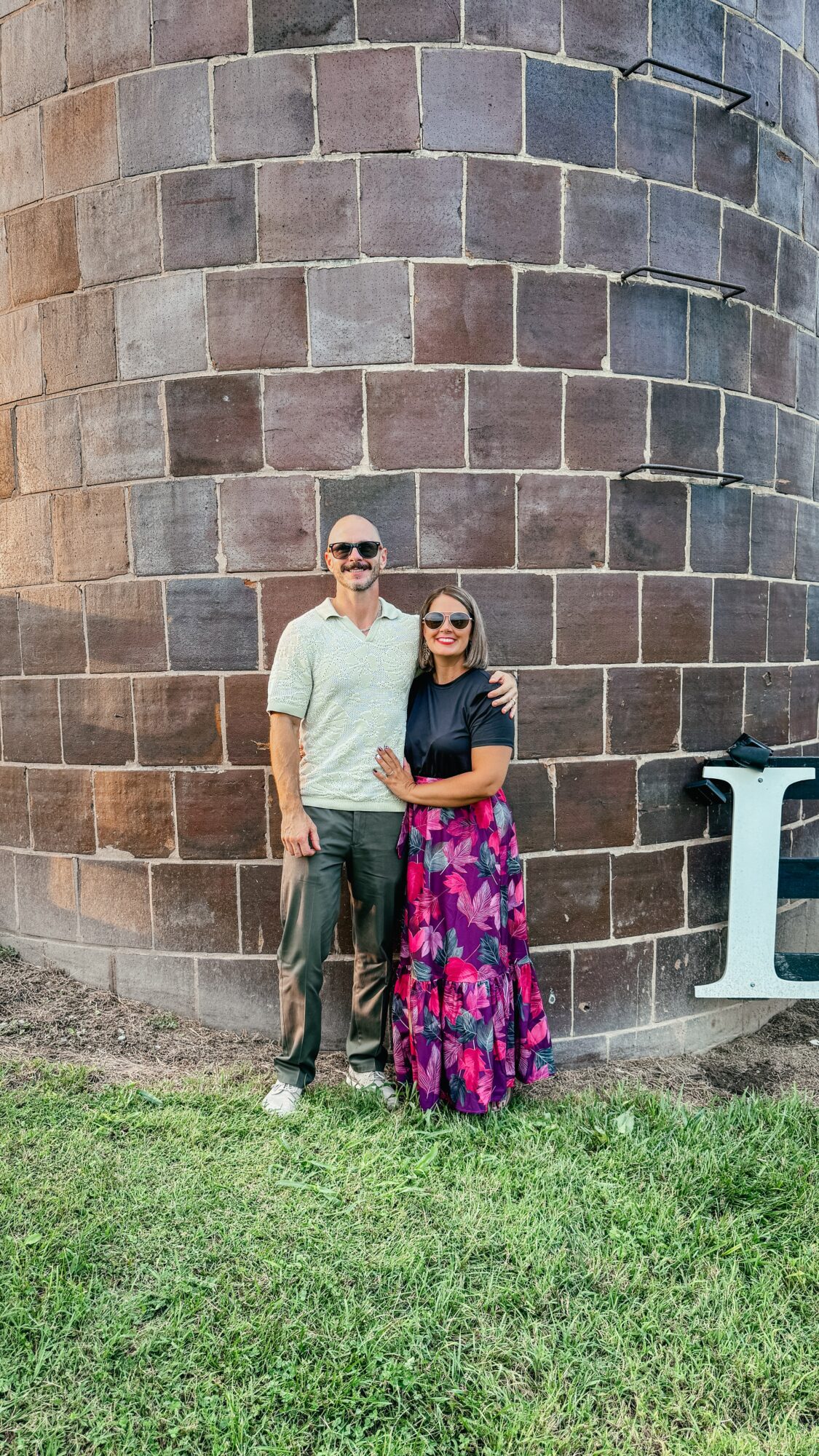 Two people stand beside a curved stone wall, with a large white letter nearby and grass in the foreground.