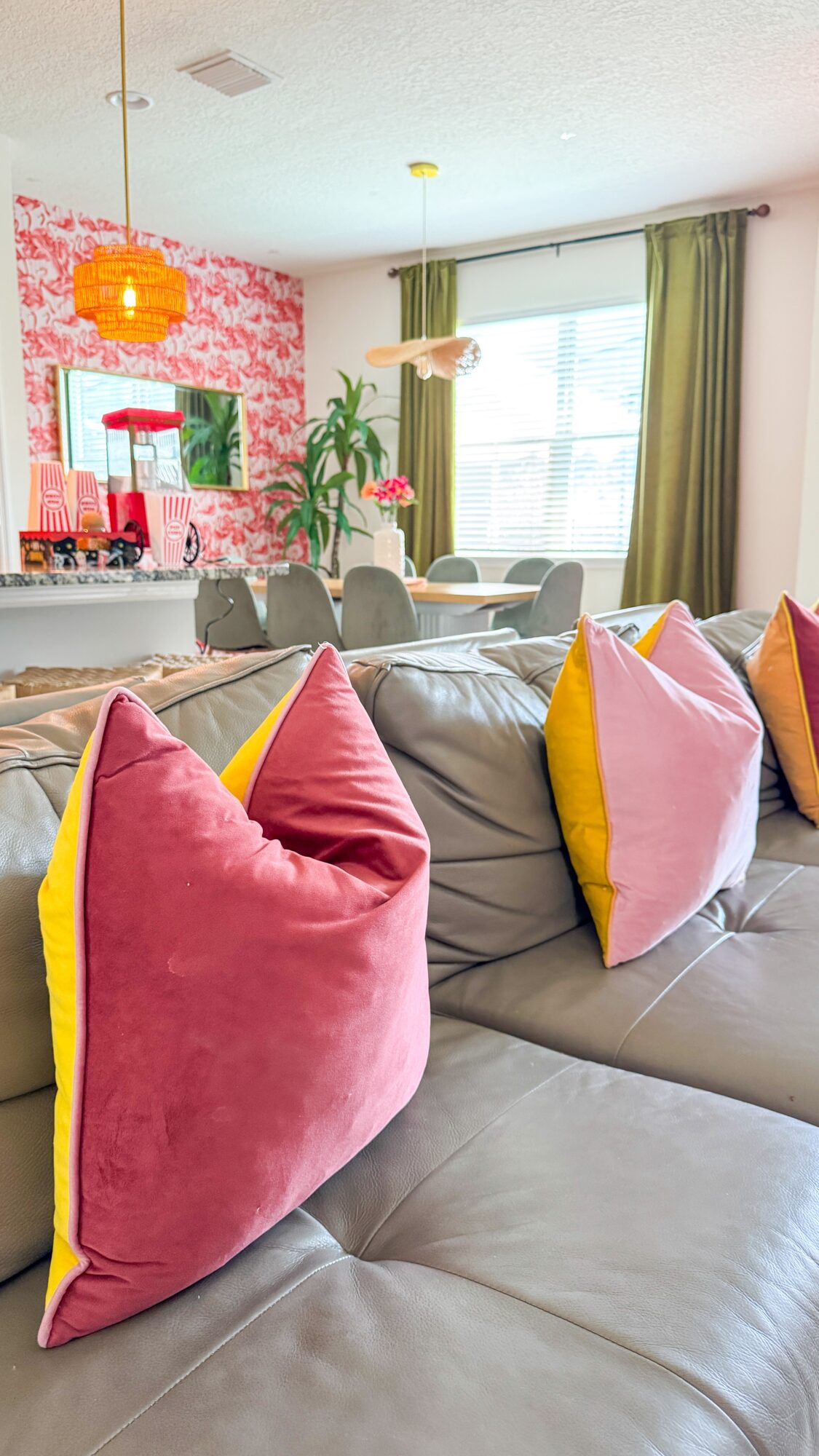 Gray sectional sofa with colorful pillows in a bright living room, with a dining area and window behind it.