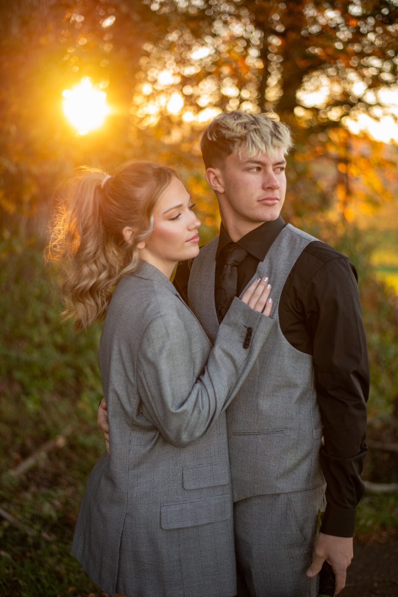 A woman and man stand together outdoors at sunset, with sunlight shining through trees behind them.
