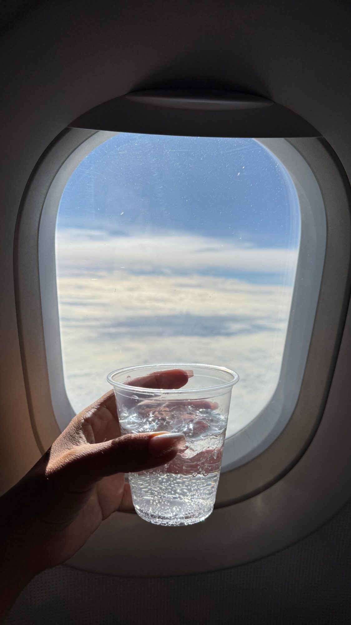 A hand holds a glass of ice water beside an airplane window showing clouds and blue sky.