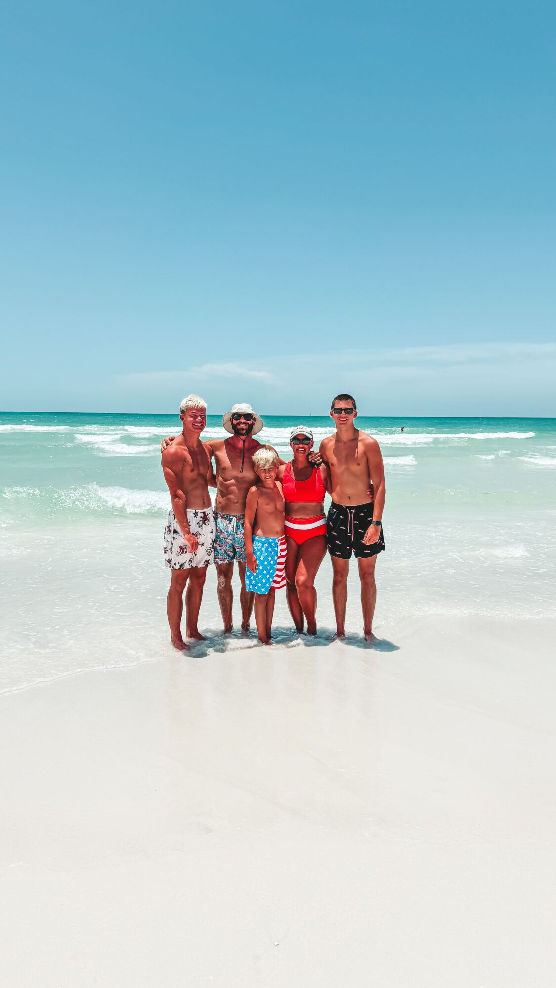 Four people stand together on a bright beach with turquoise ocean waves behind them.